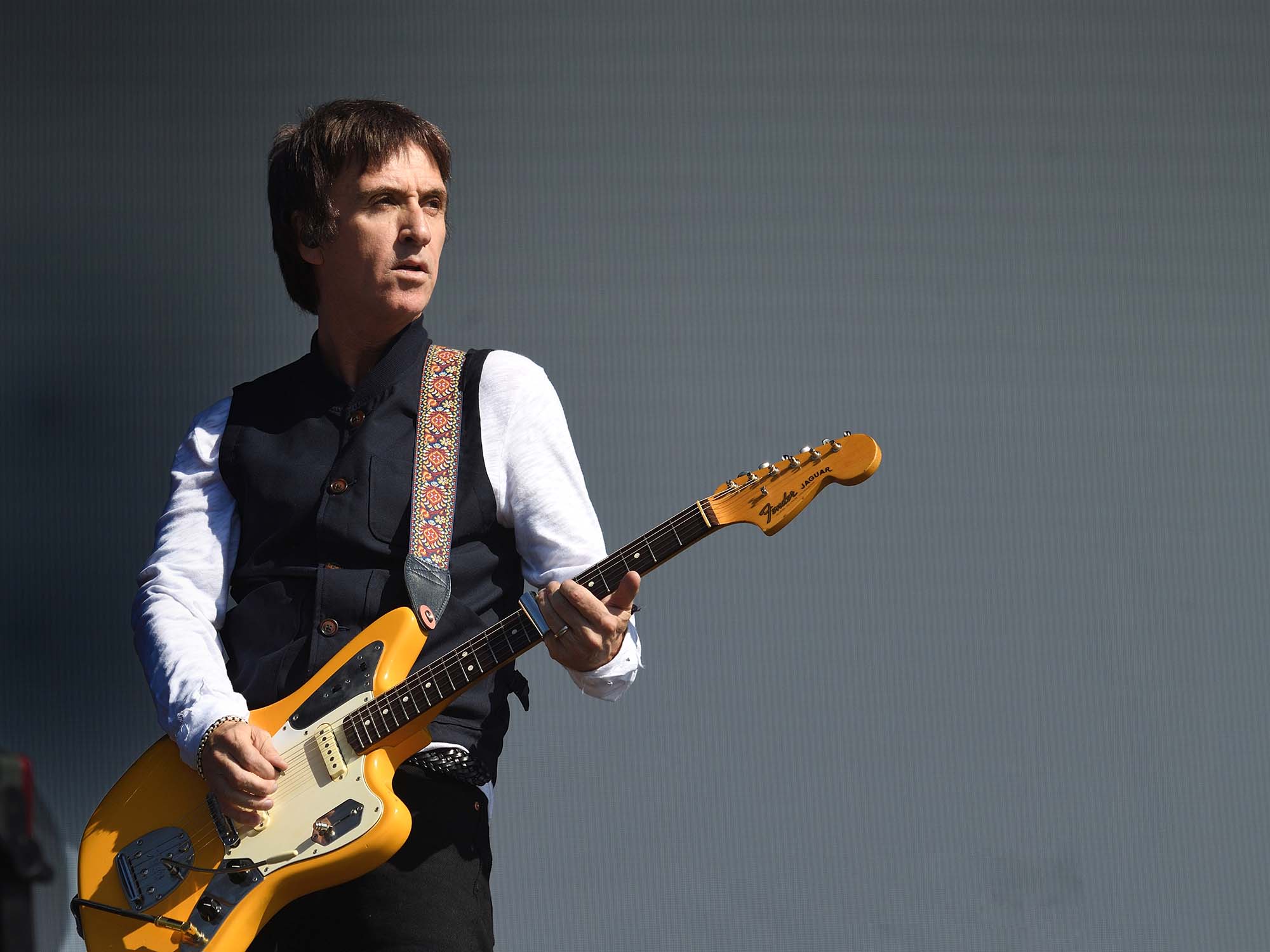 Johnny Marr playing a Jazzmaster. He is standing in front of a grey background. His guitar is a vibrant orange colour.