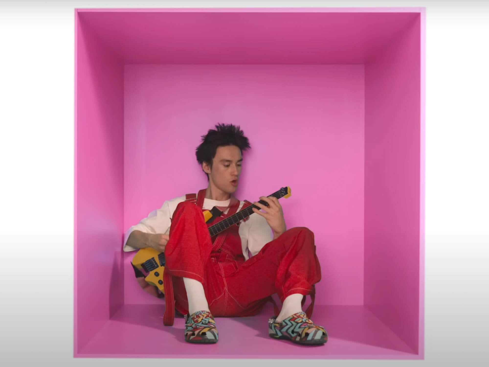 Jacob Collier in the music video for WELLLL. He's sat inside a pink box and is playing the yellow headless model