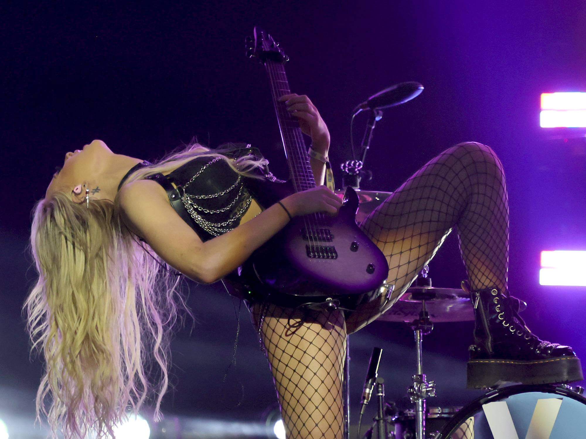 Sophie Lloyd playing guitar on stage. She has one foot placed on a drum kit and is leaning back as she is playing. She has long blonde hair which trails behind her.