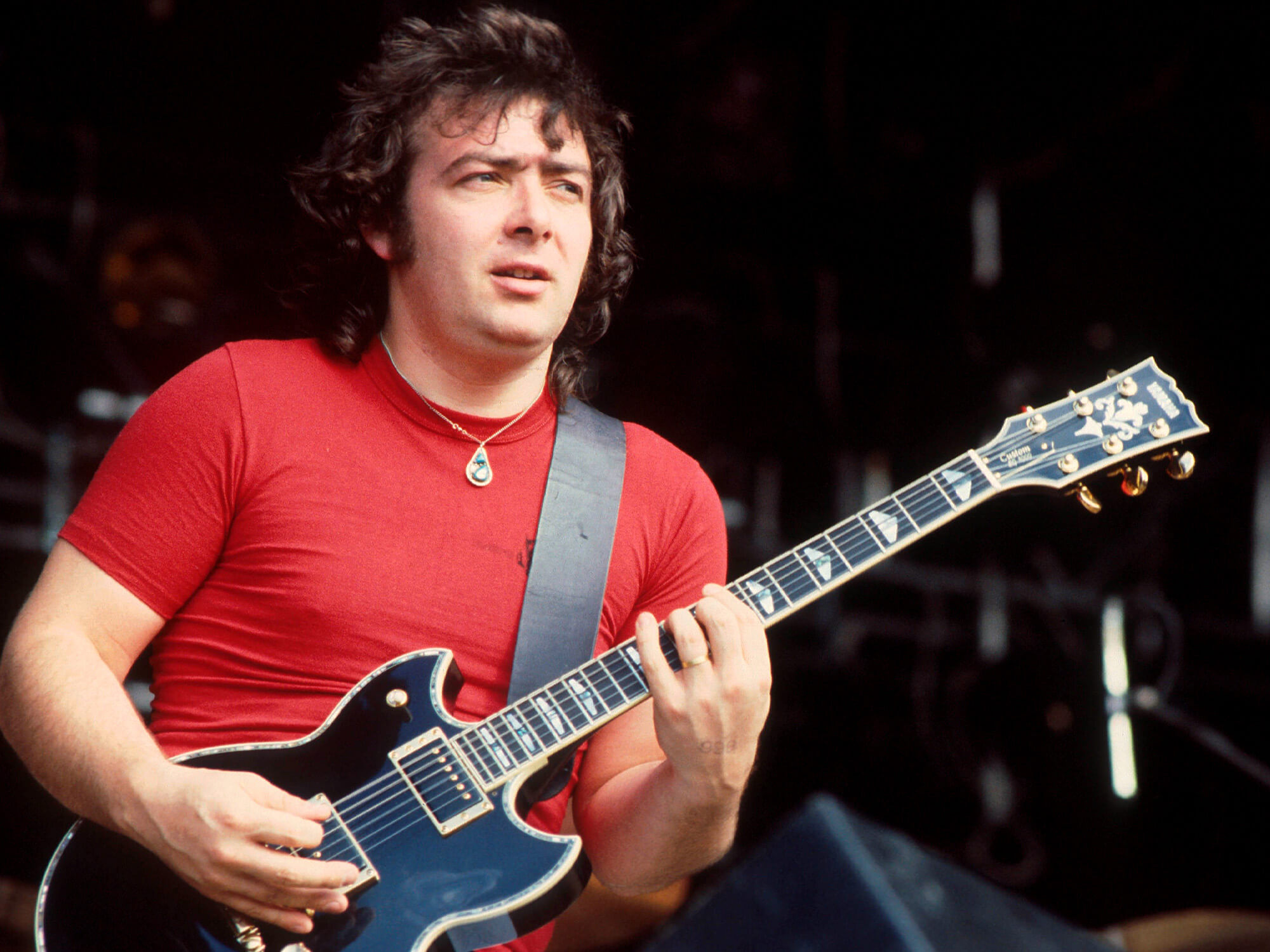 Bernie Marsden on stage in 1981, he's wearing a red shirt, playing guitar and is looking out into the crowd.