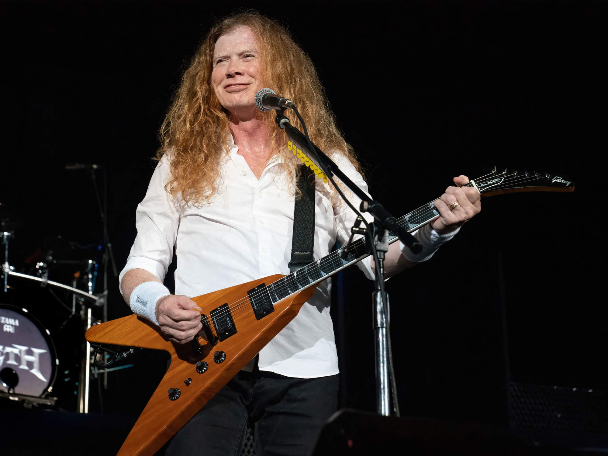 Dave Mustaine on stage. He's wearing a white shirt, is smiling, and playing a Gibson V guitar