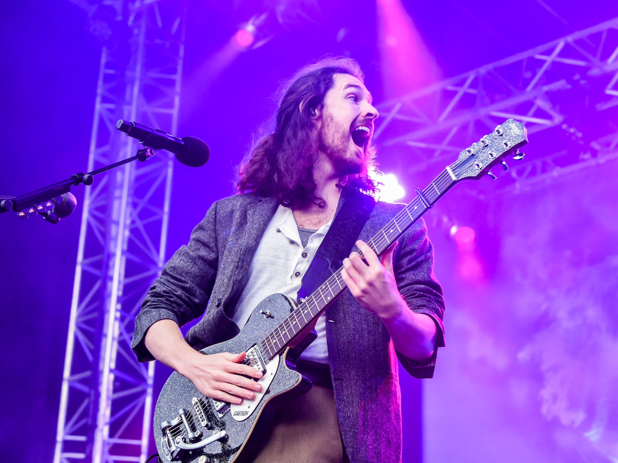 Hozier playing a black sparkle Gretsch guitar on stage. He's smiling and stands on stage amongst purple lighting