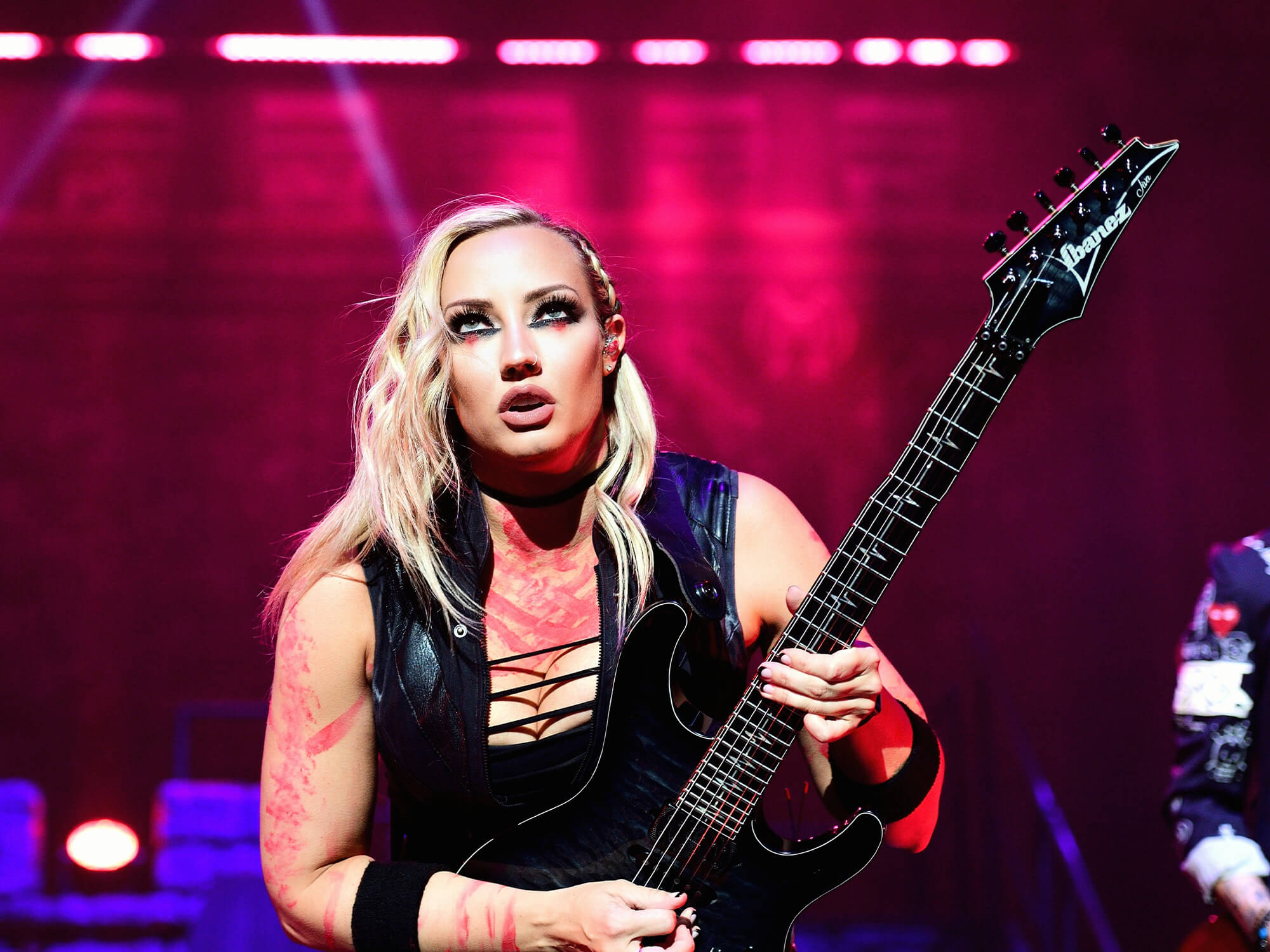 Nita Strauss on stage, she is crouched and playing her Ibanez electric guitar. She's looking forwards out into the audience.