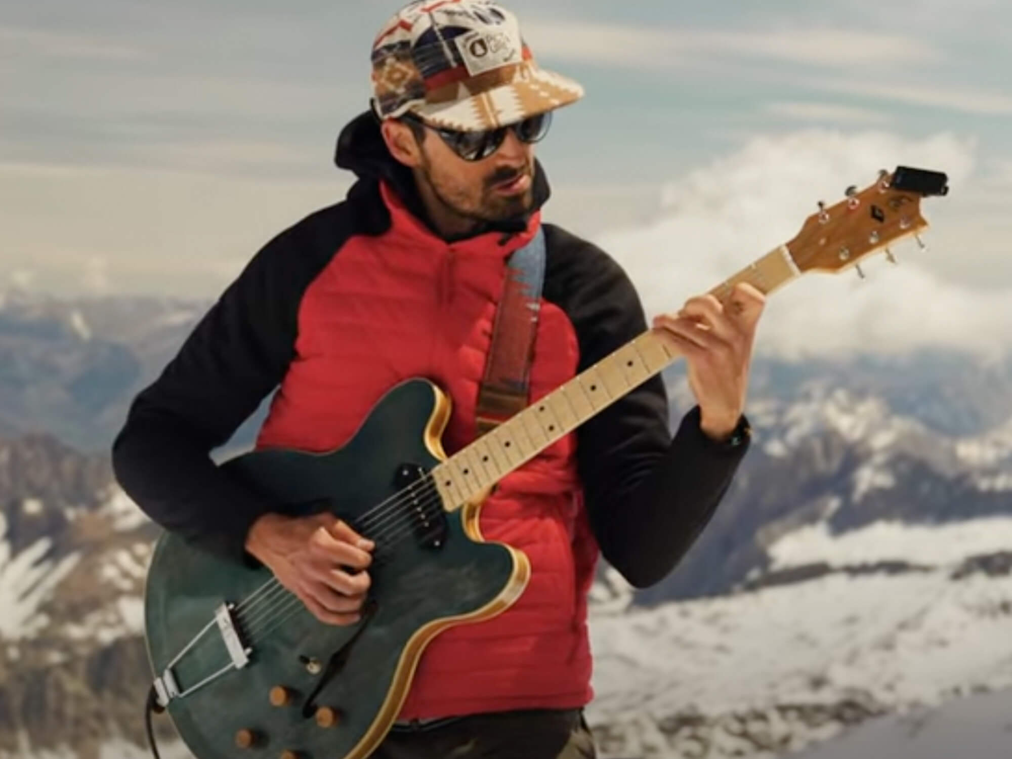 Lopez performing his original song on a blue custom semi-hollow guitar. He's wearing a red coat and the mountain range is behind him.