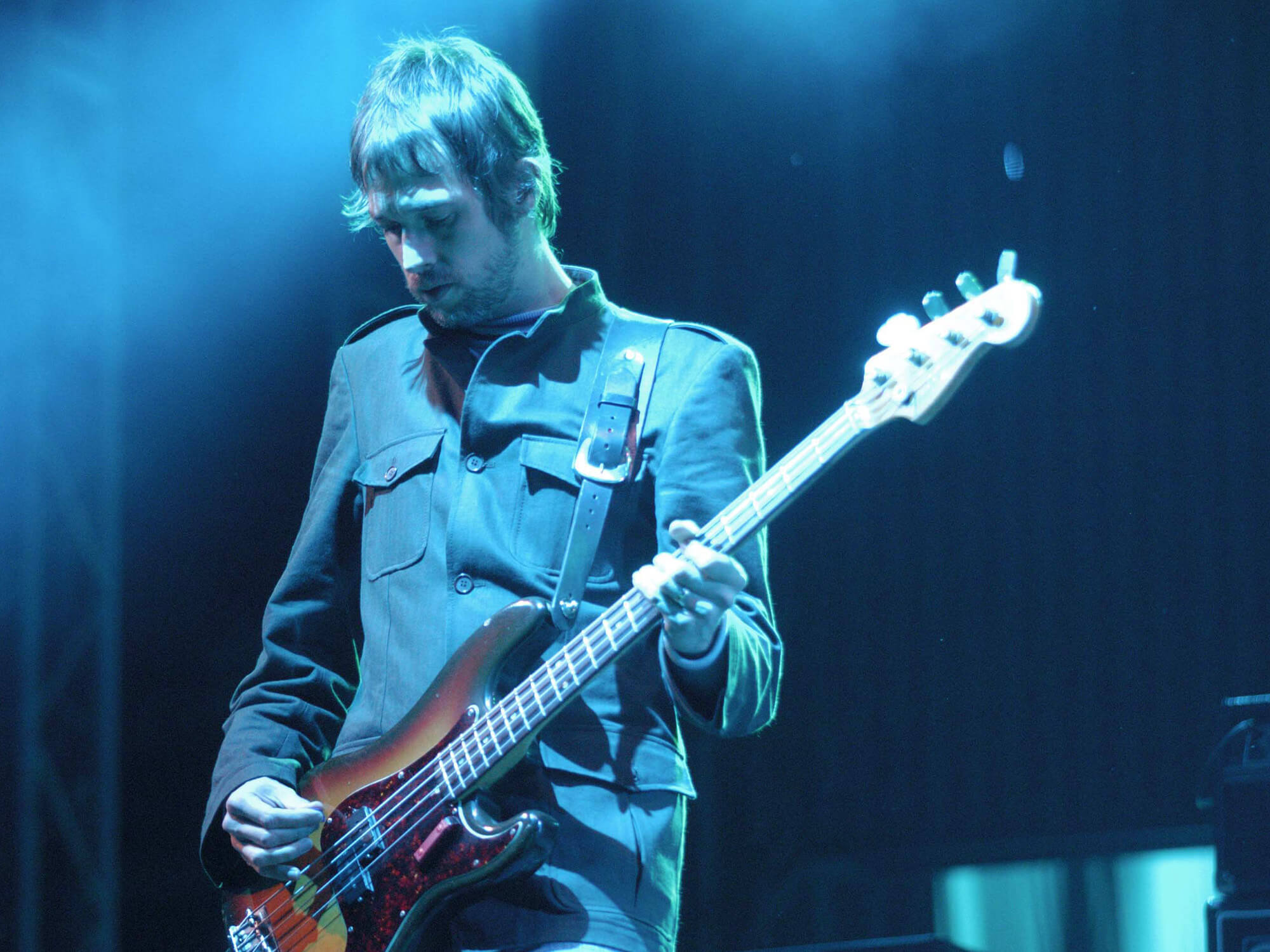 Andy Bell playing bass with Oasis. He's looking down at his instrument and stands under blue lighting.
