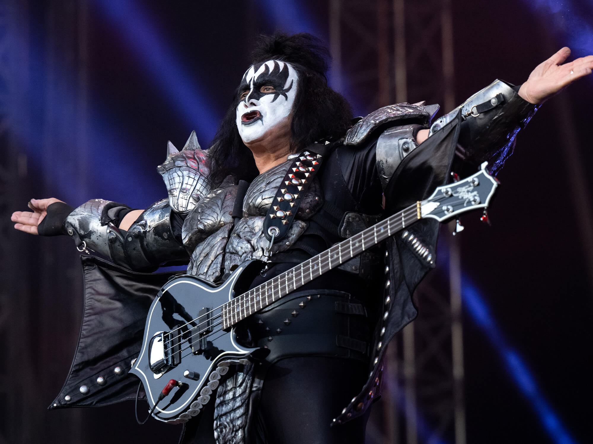 Gene Simmons on stage with both arms out to the side, and his bass guitar at his torso