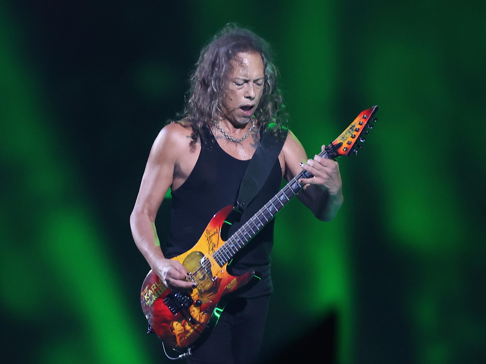 Kirk Hammett playing guitar on stage. He stands before green lighting and is looking down at his instrument.
