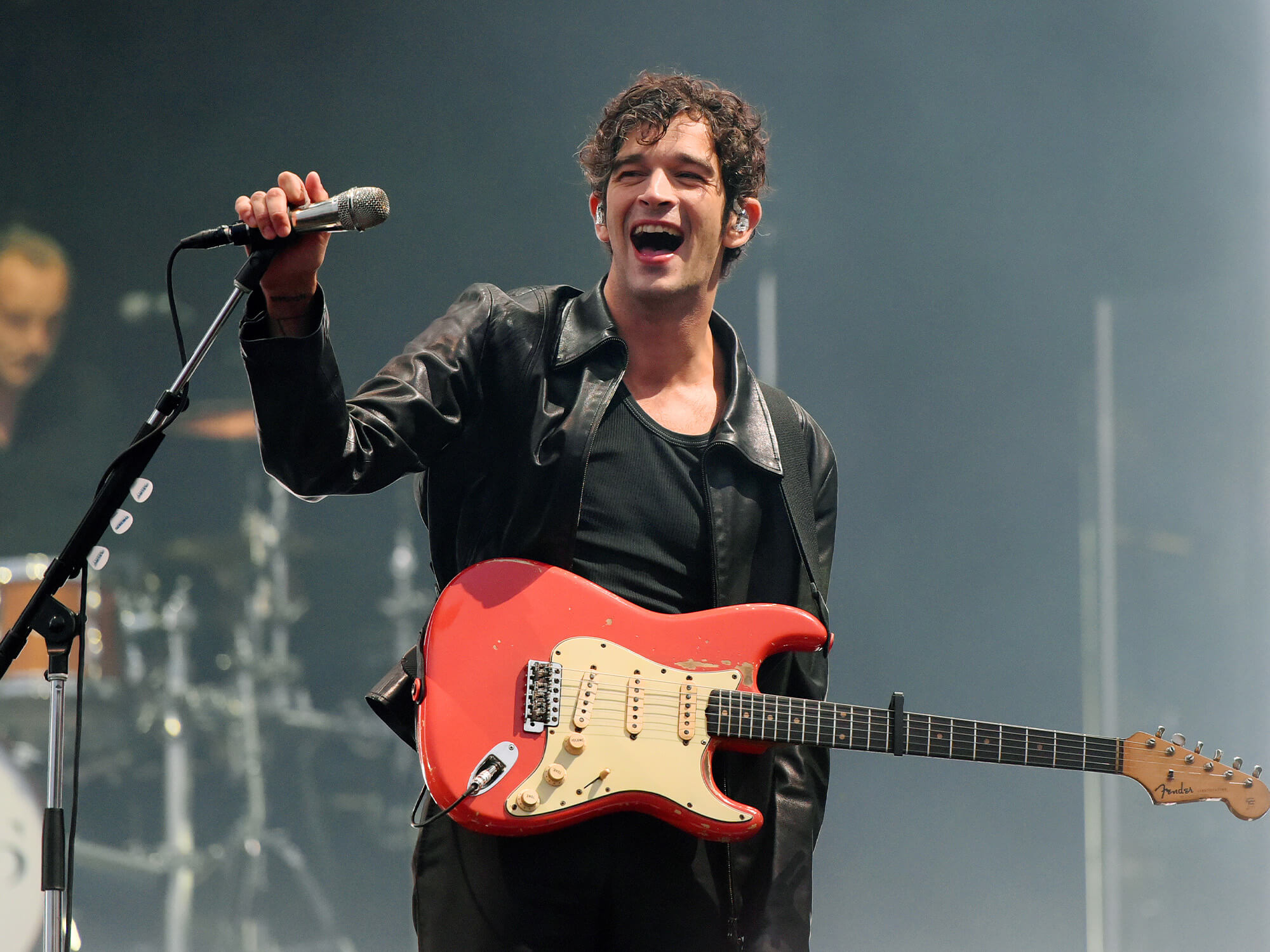 Matty Healy gripping a mic stand with one hand and smiling open mouthed whilst on stage. He has a red Fender Strat hanging from his torso.