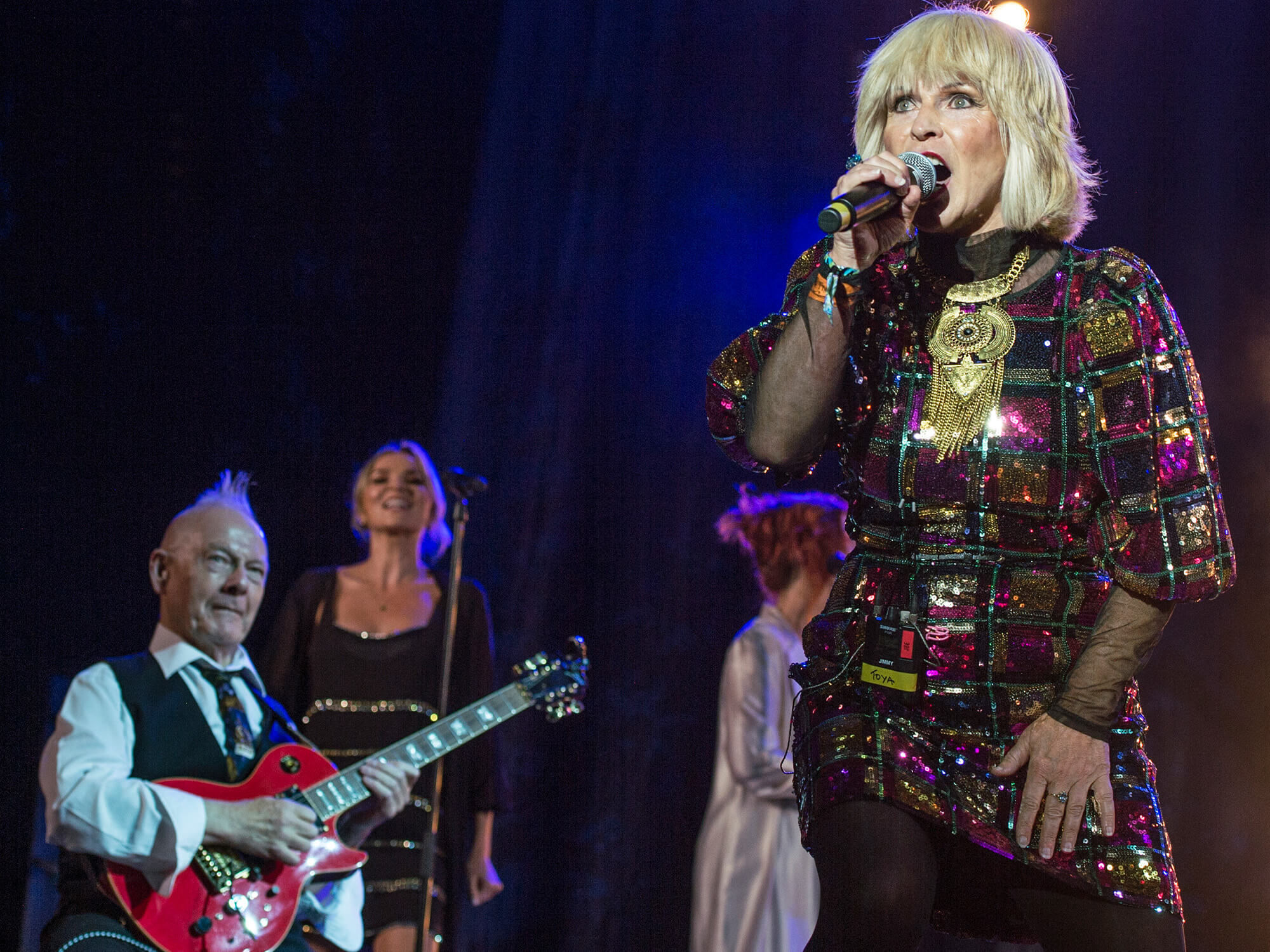 Toyah and Robert on stage. Robert is sat playing guitar whilst Toyah sings into a microphone wearing a sparkling dress.