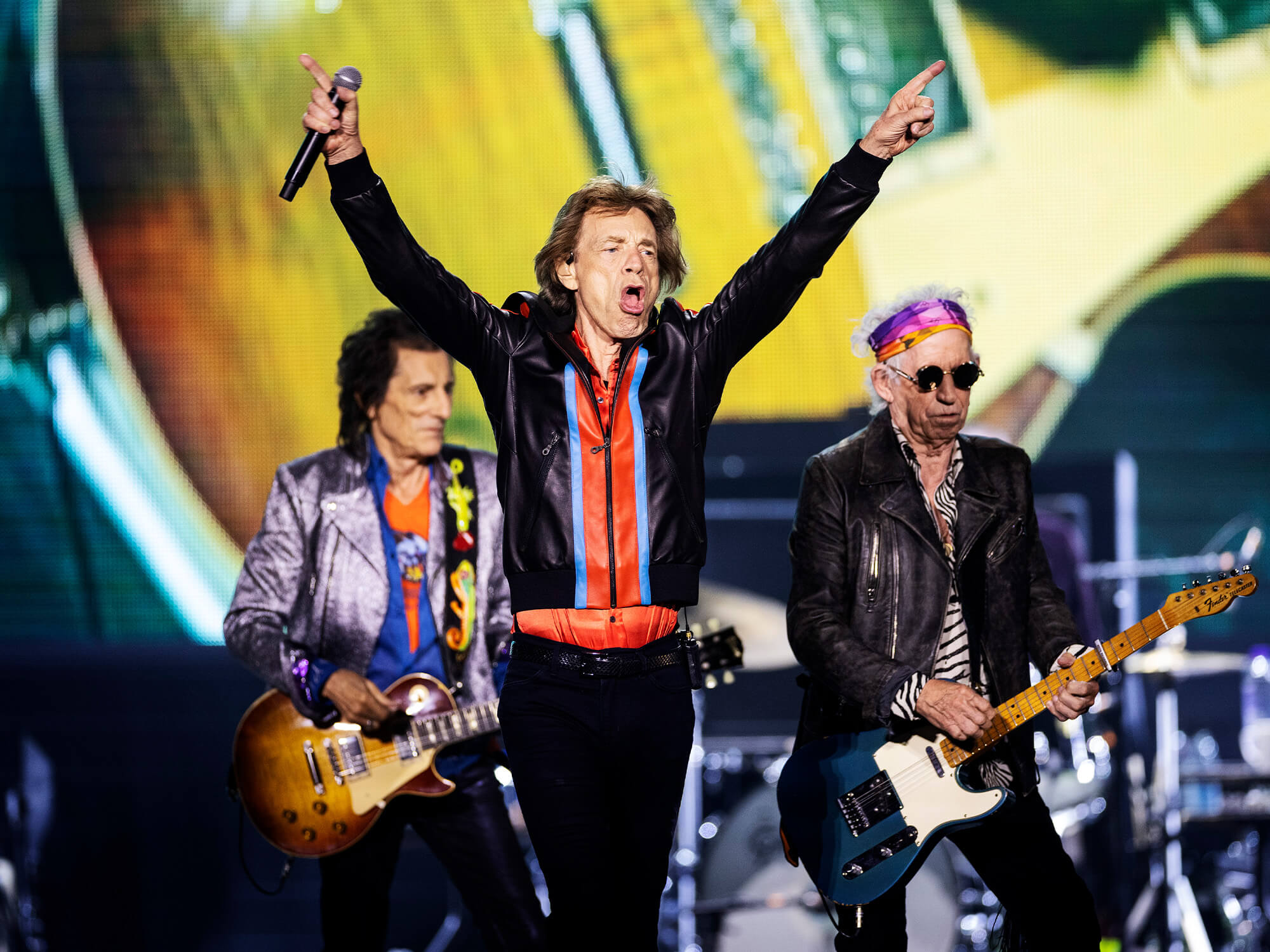 The Rolling Stones on stage. Mick Jagger has both hands in the air with an open-mouthed expression.