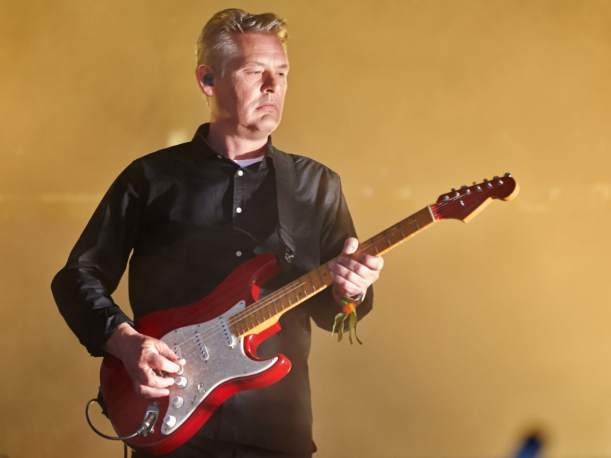 Angelo Bruschini on stage in 2014. He is playing a red Strat guitar and has a plain expression on his face.