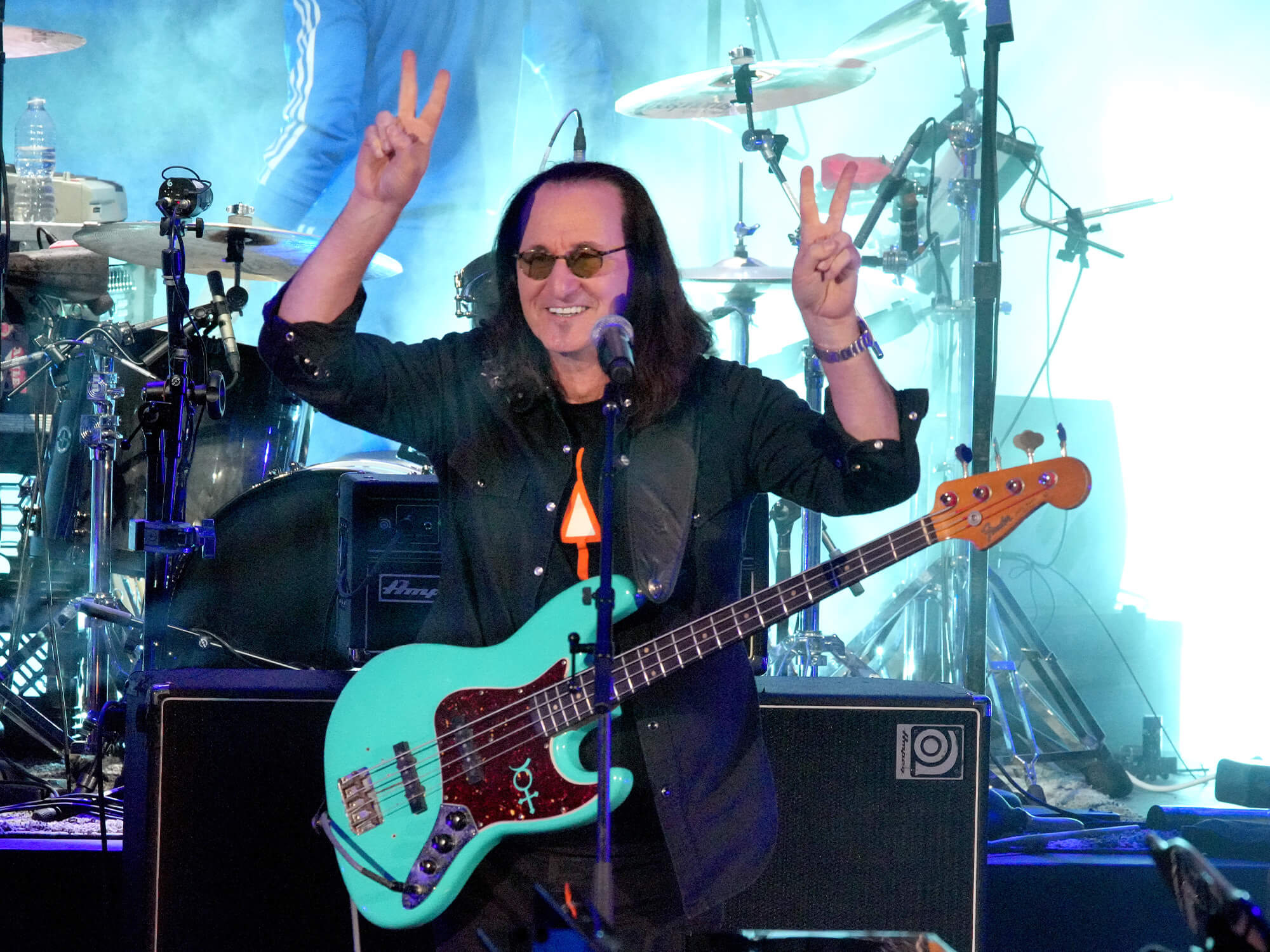 Geddy Lee on stage. He has a bass guitar hanging at his torso and is smiling and holding up two peace signs with his hands.