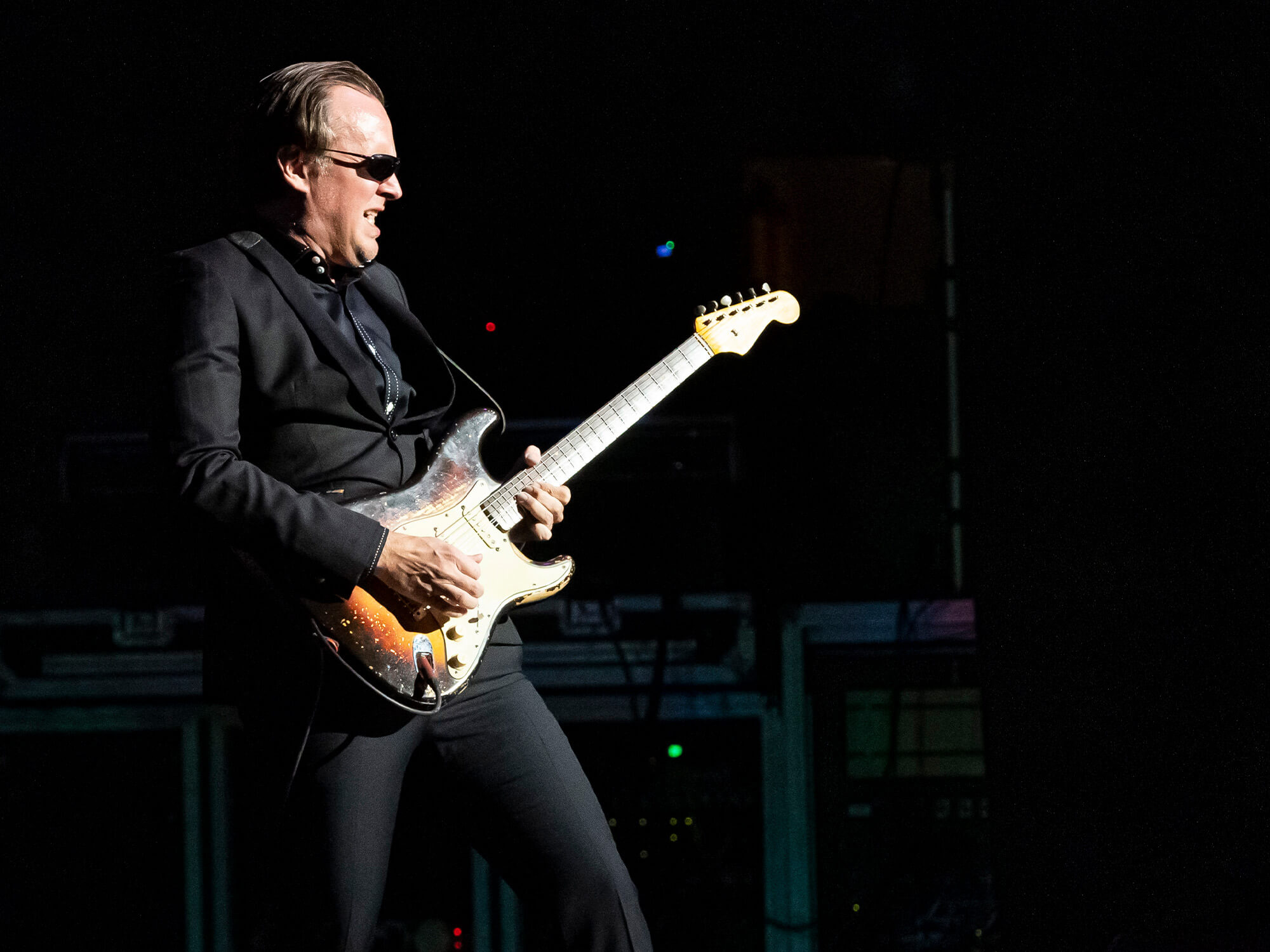 Joe Bonamassa on stage playing a Strat. He stands before a black backdrop and wears his famous suit and sunglasses