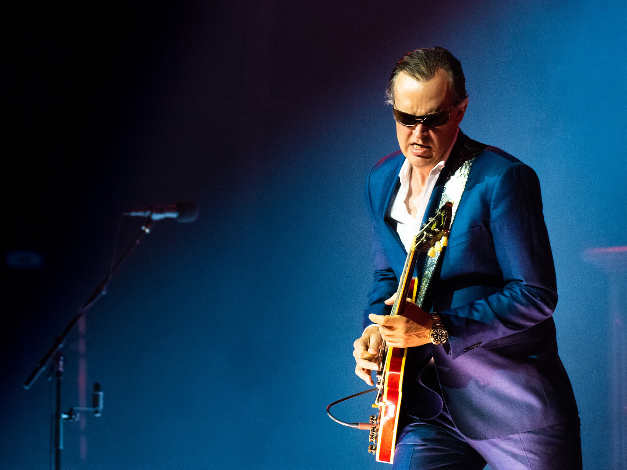 Joe Bonamassa wearing sunglasses and a suit, playing guitar on stage