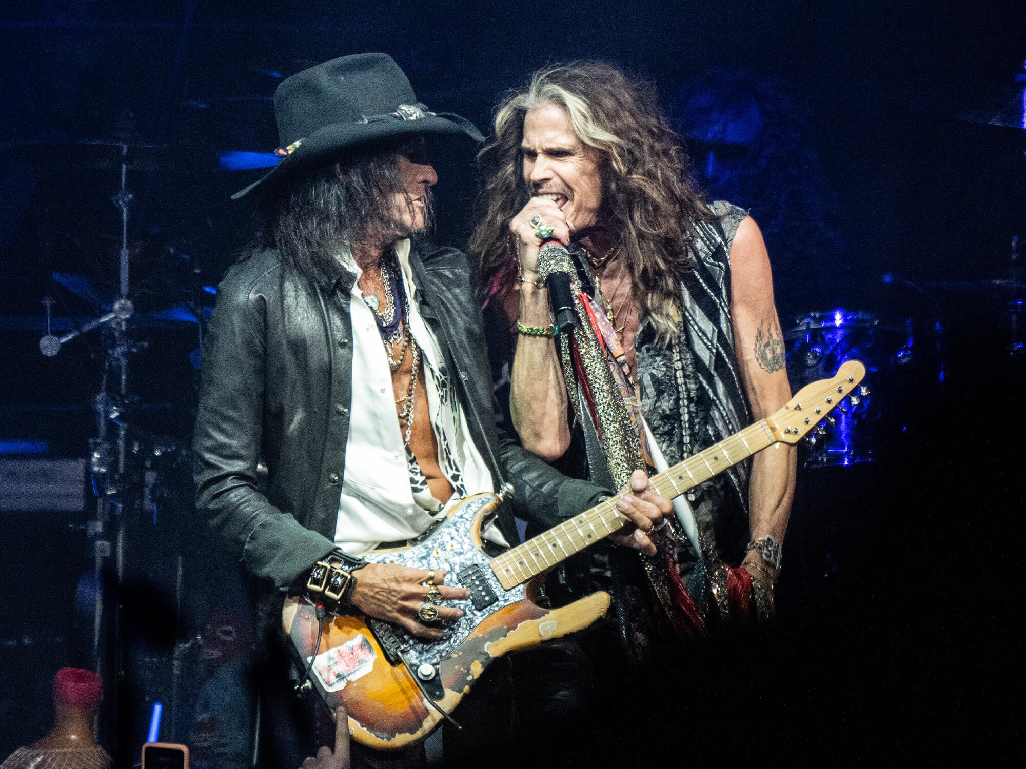 Joe Perry playing guitar next to vocalist Steven Tyler who is singing into a mic and looking back at him