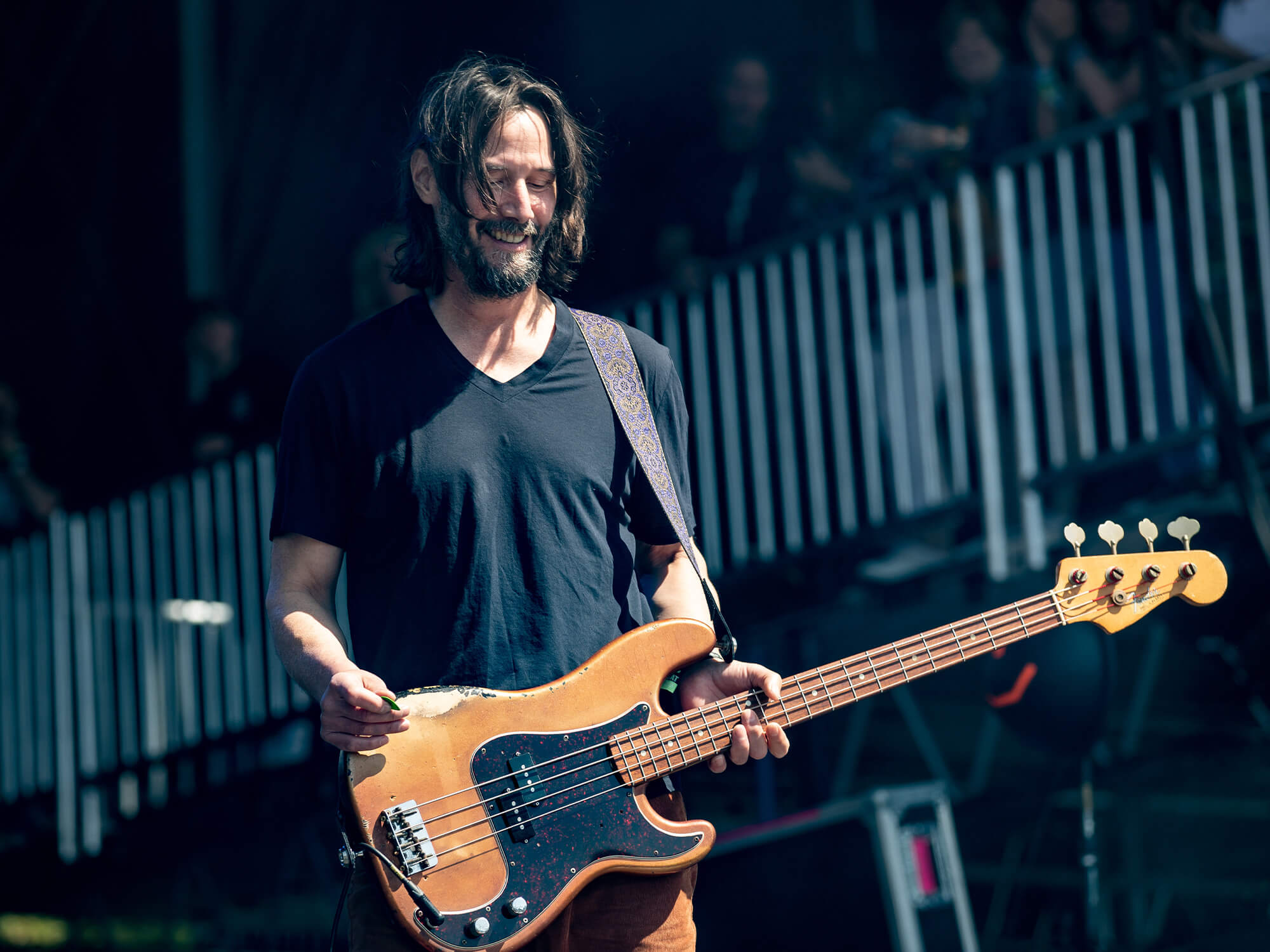 Keanu Reeves on stage holding his bass guitar. He's looking down at it at smiling.