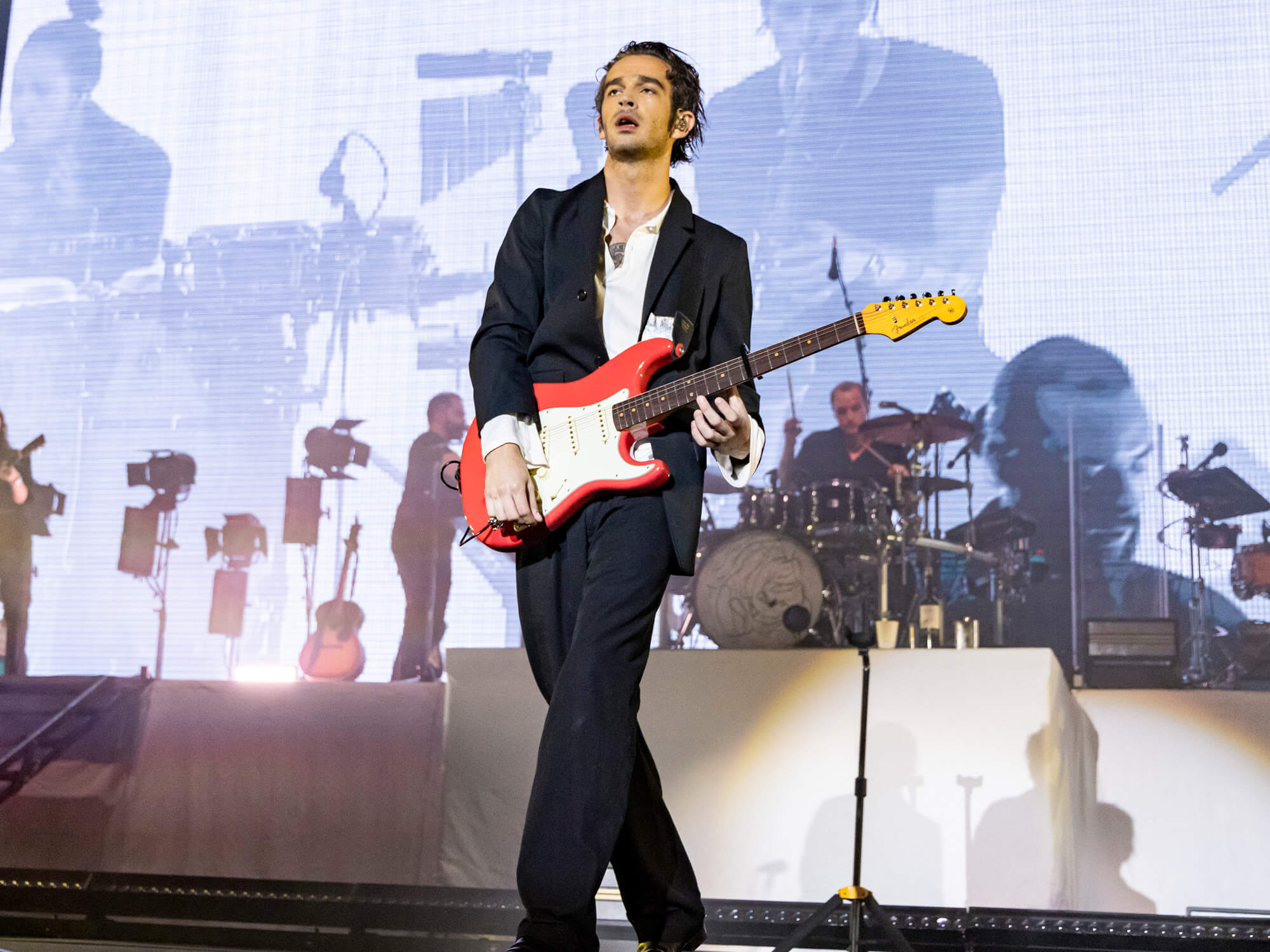 Matty Healy on stage wearing a suit and playing a red Stratocaster guitar