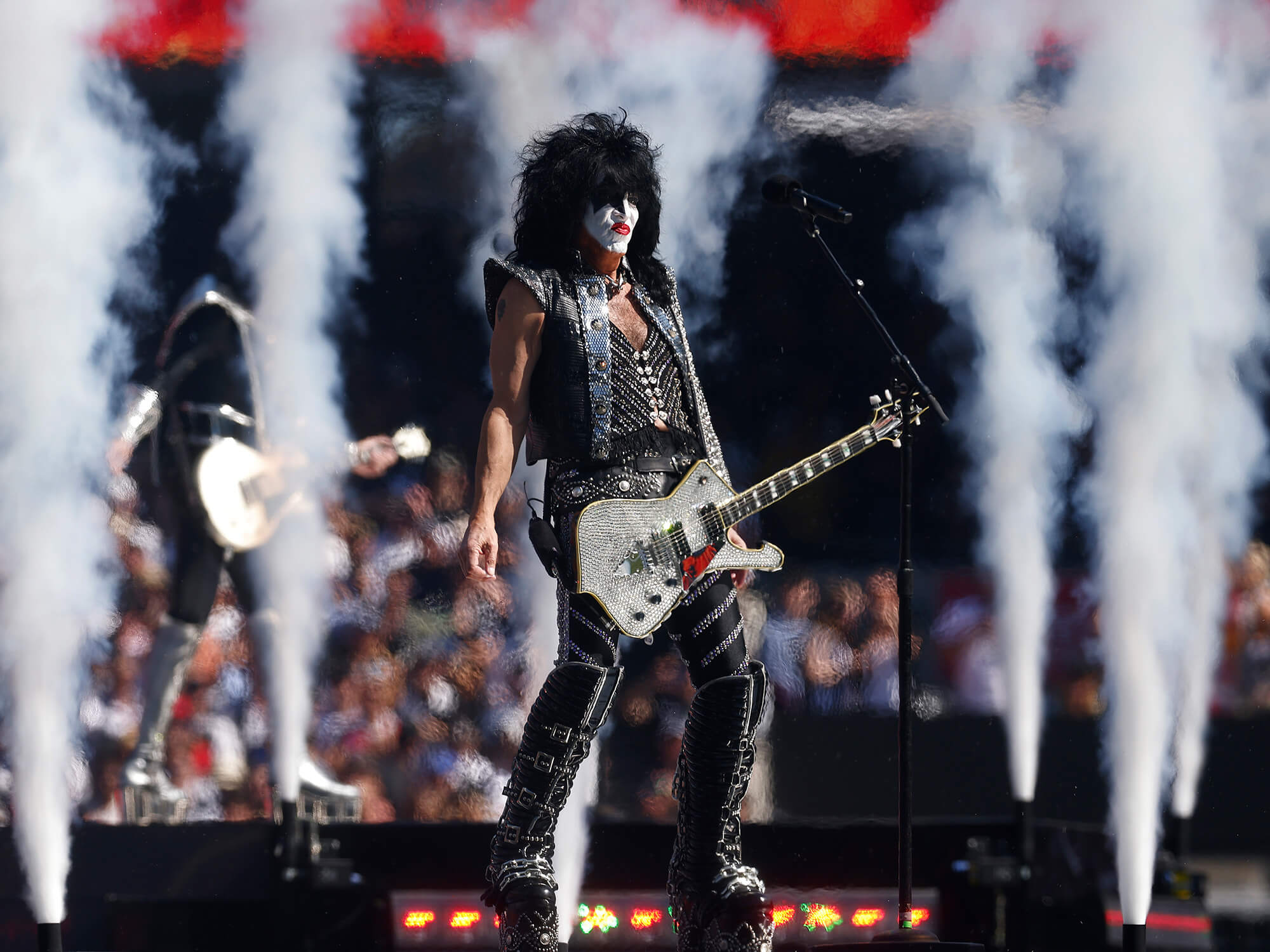 Paul Stanley on stage with his bejewelled guitar. He wears his signature white face paint, red lips and black eye make up. Smoke machines are going off behind him.