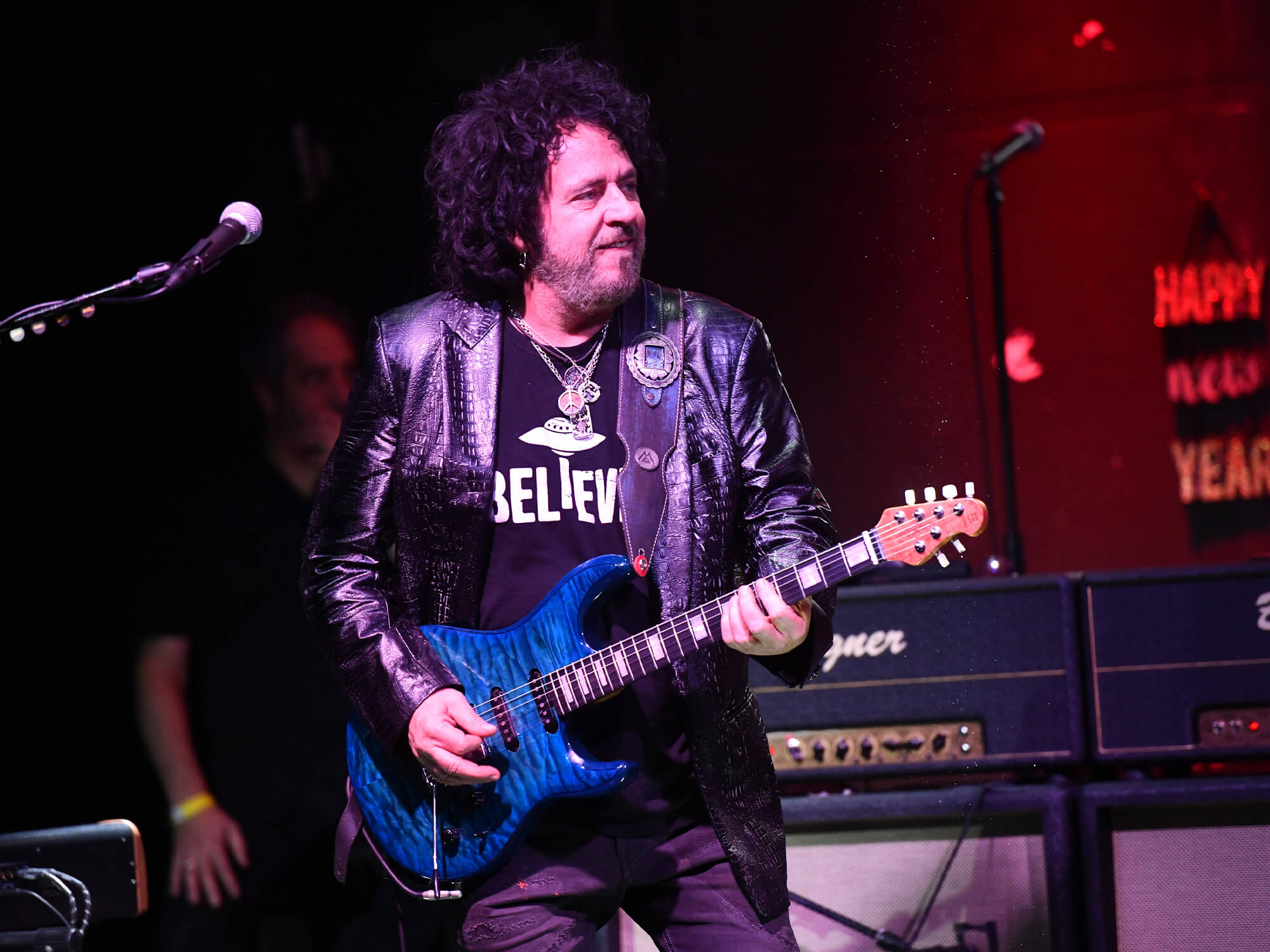 Steve Lukather on stage playing a blue Ernie Ball guitar. He is smiling and looking to his left.