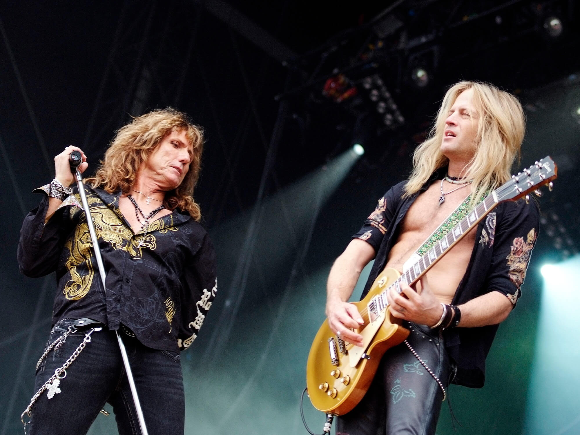 David Coverdale holding a microphone attached to a stand. He's looking at Doug Aldrich who is playing a Les Paul guitar