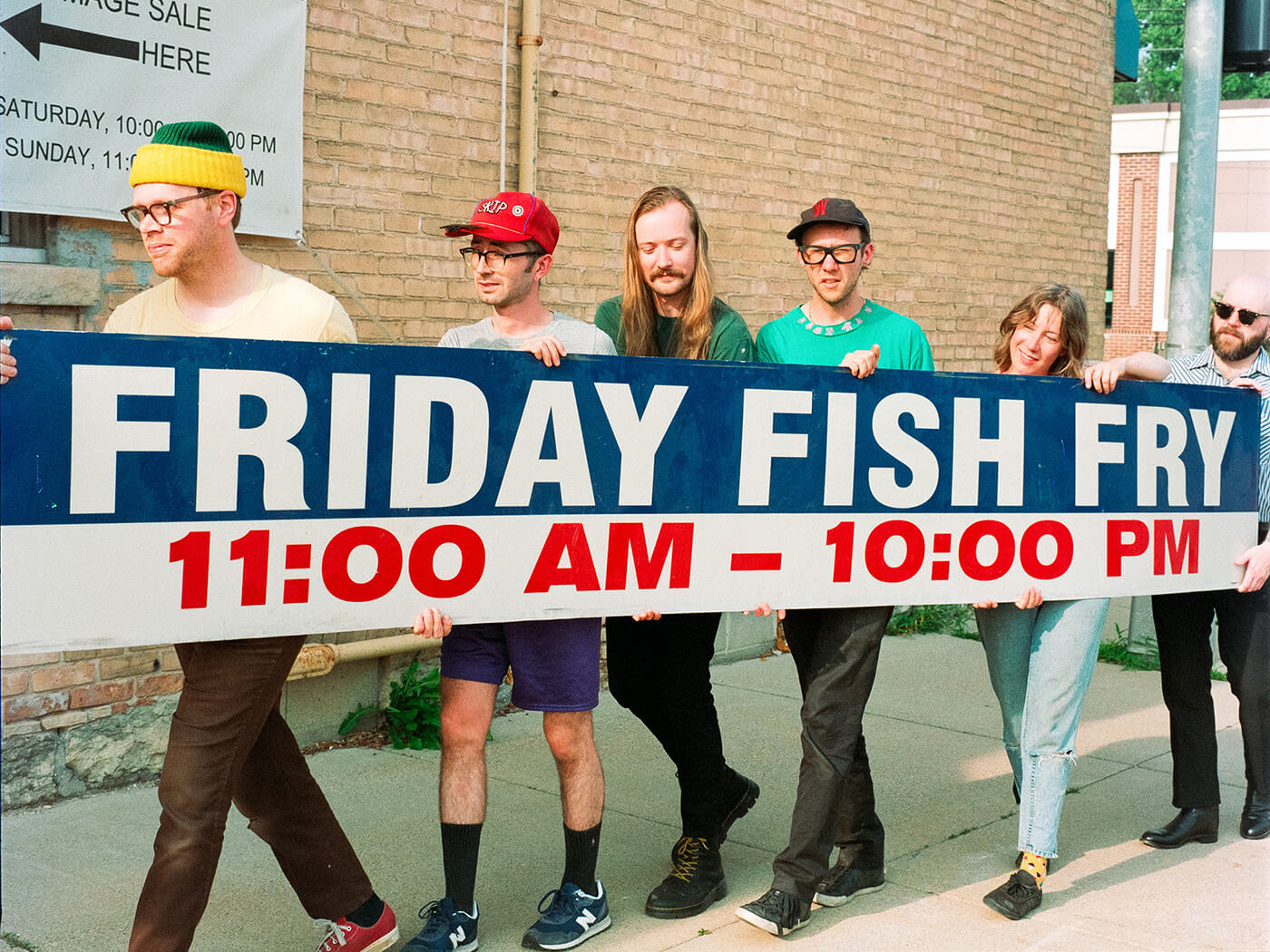 The band Dusk holding up a sign that says ‘Friday Fish Fry 11:00 AM - 10:00 PM’, photo by Billy Hintz