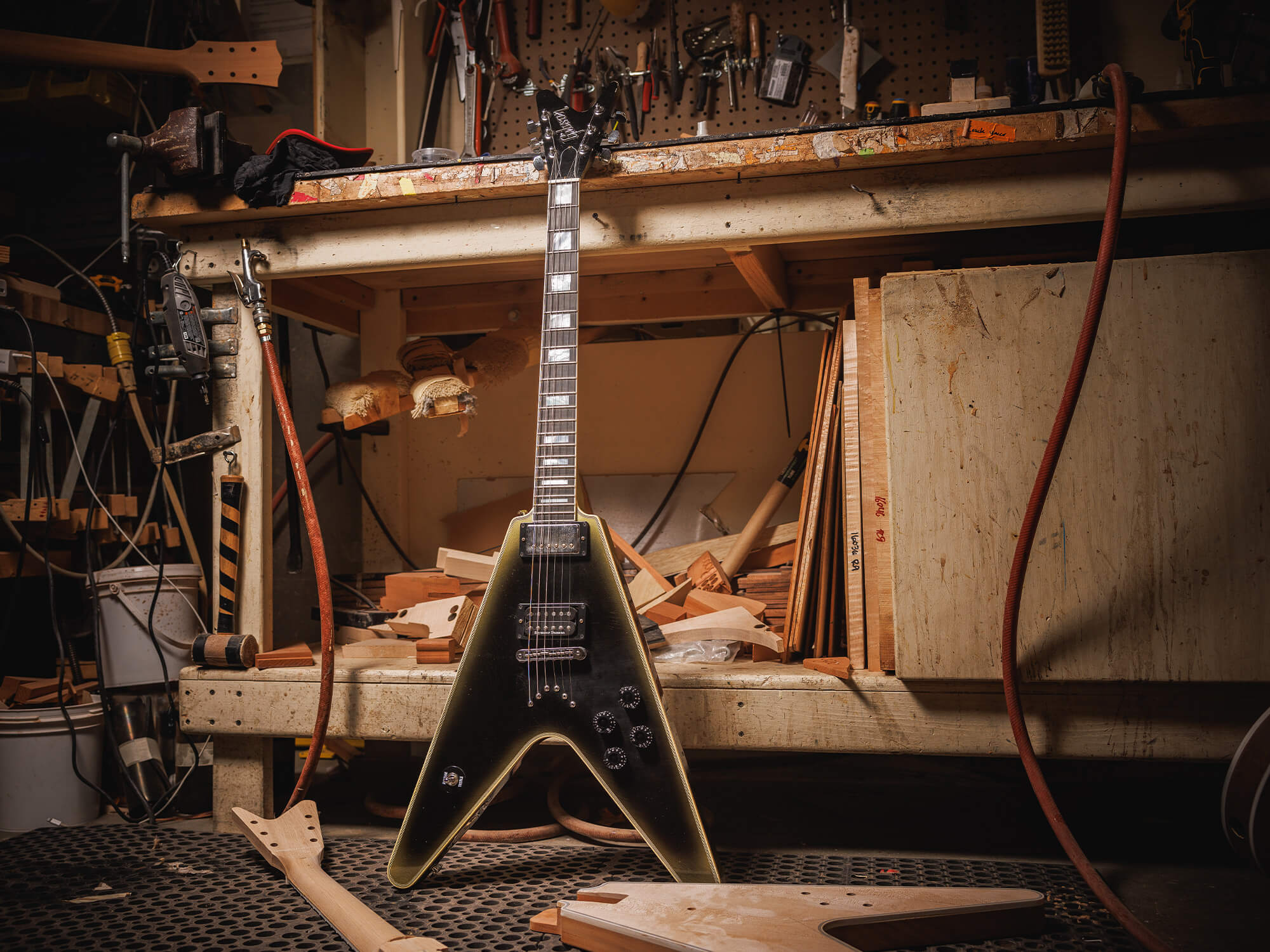 The Adam Jones Flying V. It is in a workshop environment, leaning against a workbench. It is a V-shaped electric guitar that looks rustic and has a dark body with golden edges.