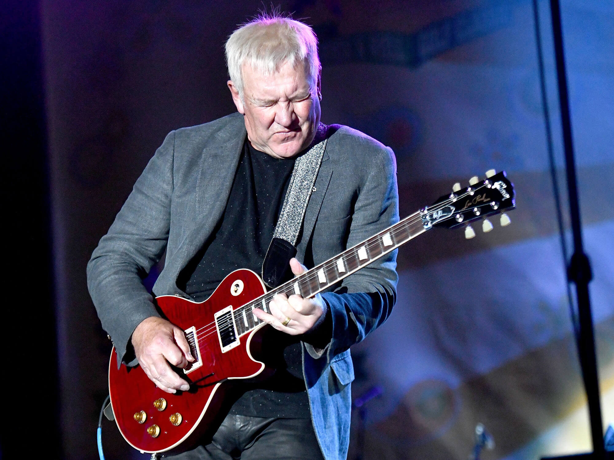 Alex Lifeson playing a red Les Paul. He has his eyes closed and is playing with a passionate expression on his face.