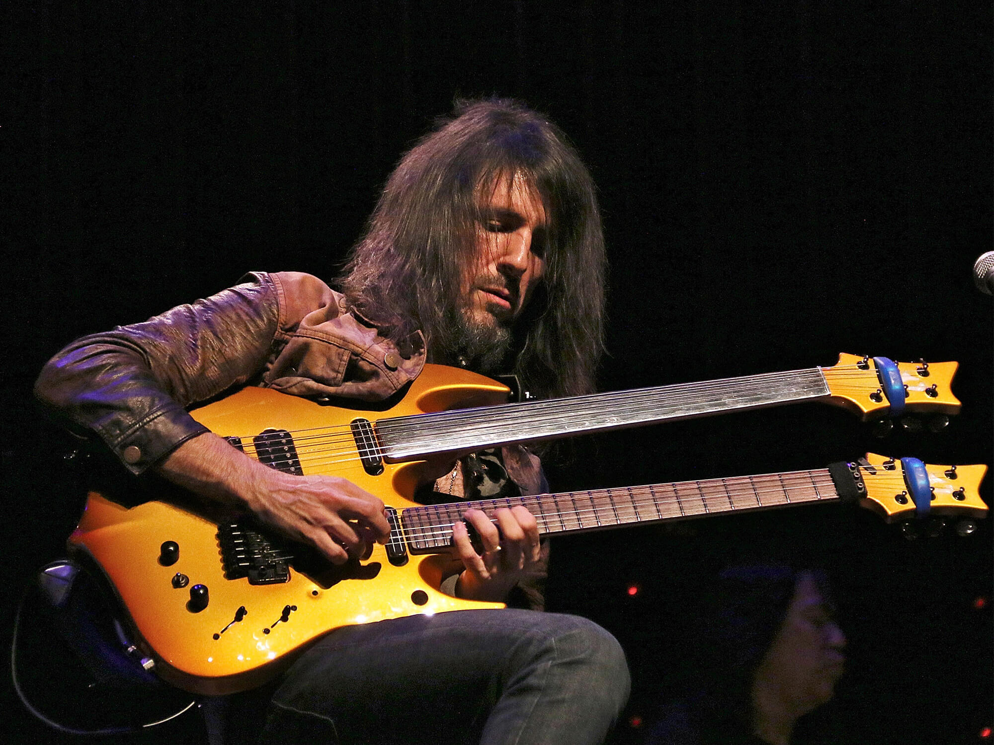 Bumblefoot playing a yellow double neck guitar. He has his fingers positioned high up on the frets of the lower guitar and is looking down as he is playing.