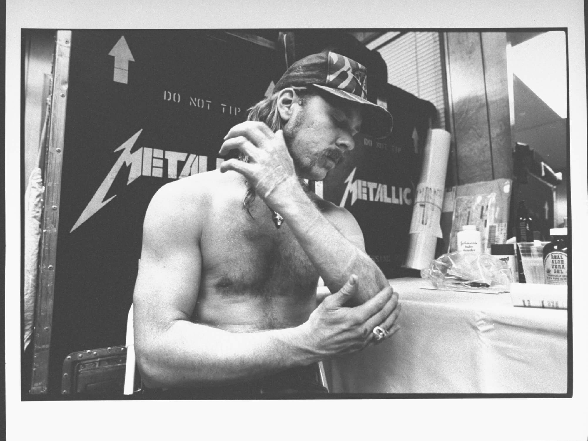 James Hetfield taking care of his burns following pyro incident. He is sitting and rubbing a gel on his arm. The photo is in black and white.