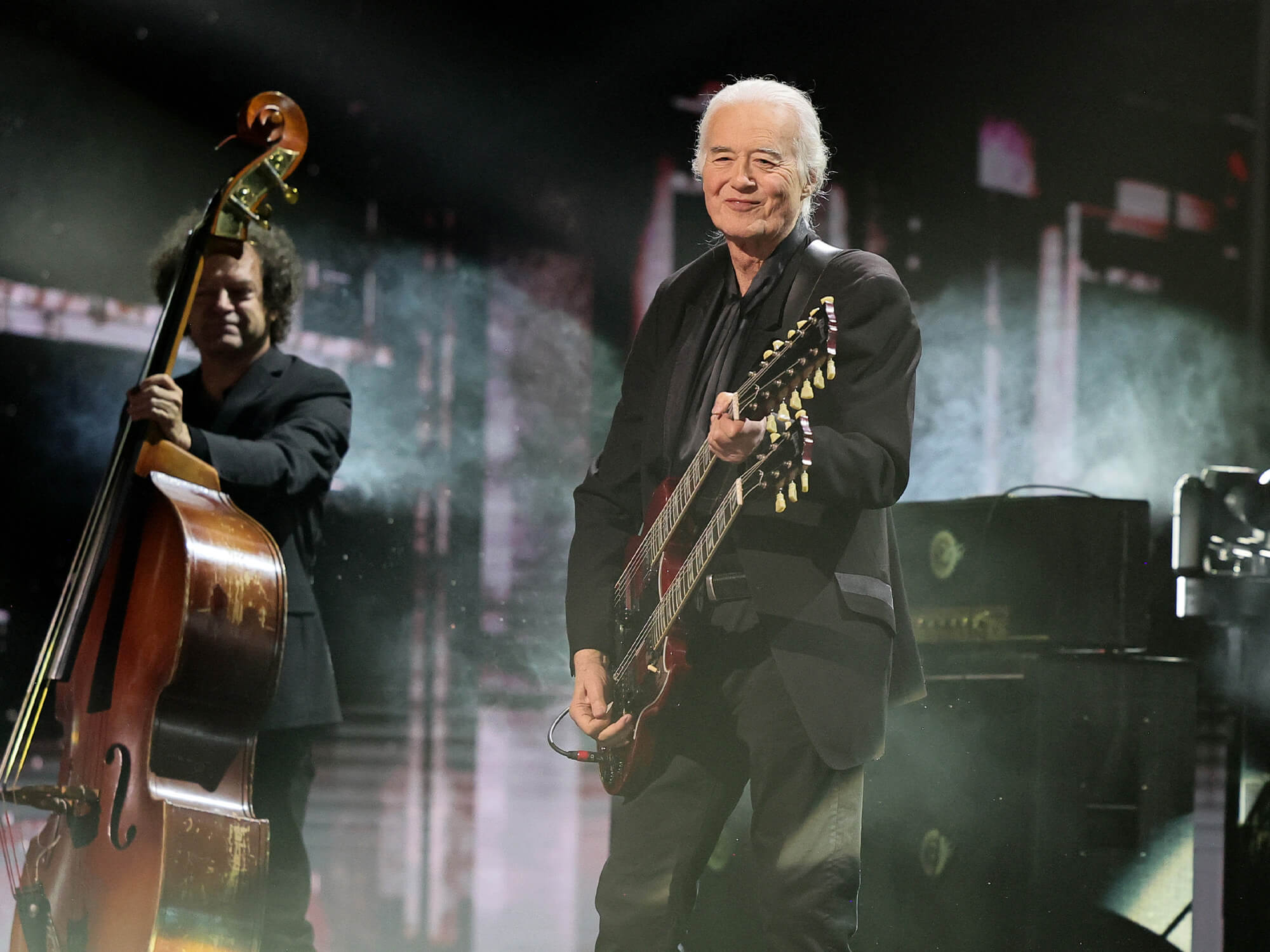 Jimmy Page smiling on stage at the Rock Hall induction performance. He is holding his double neck guitar.