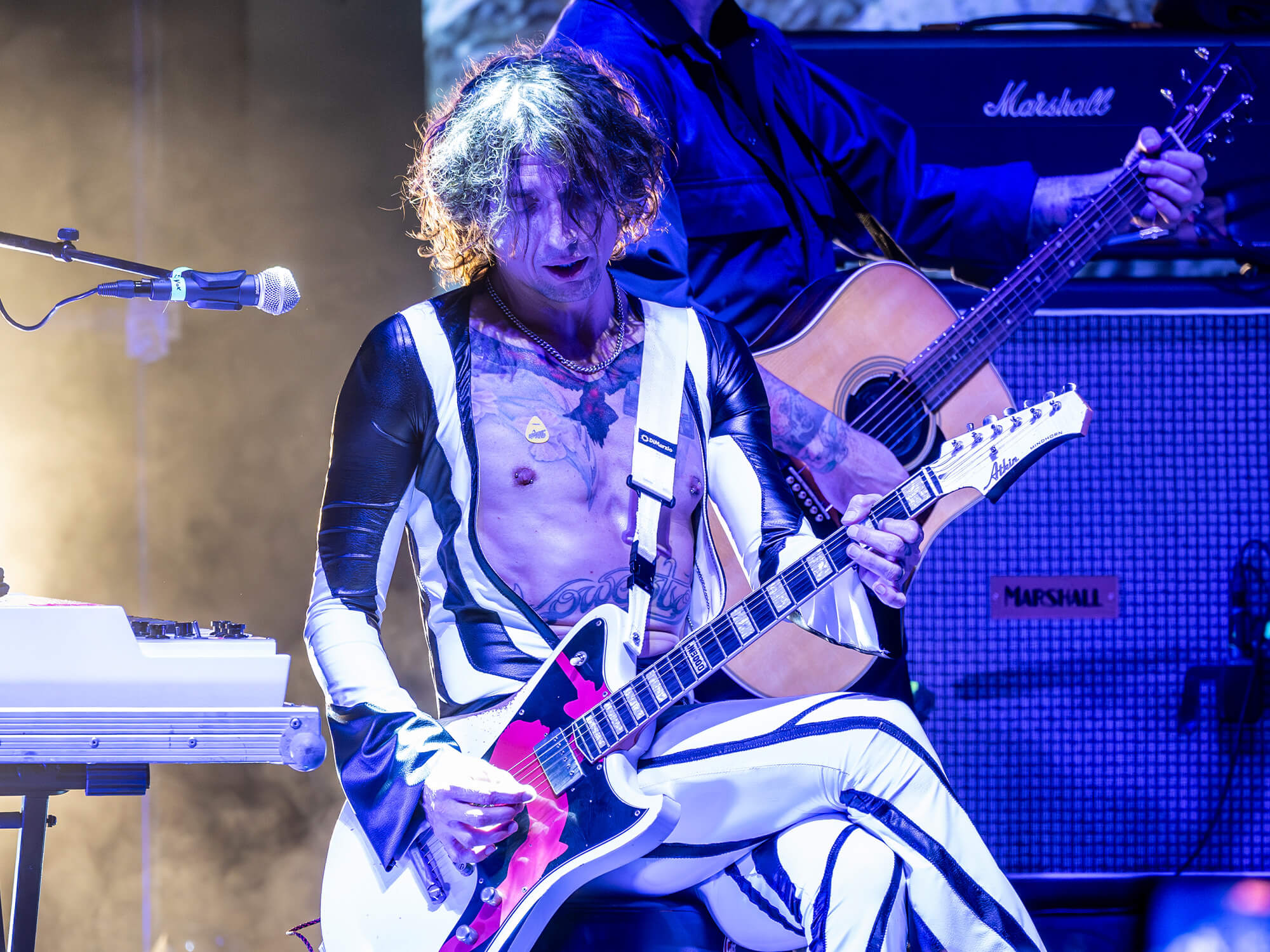 Justin Hawkins on stage. He is playing guitar and sitting with one leg crossed over the other. The lighting around him is blue, and he is wearing a striped one-piece outfit with flared sleeves and trouser legs.