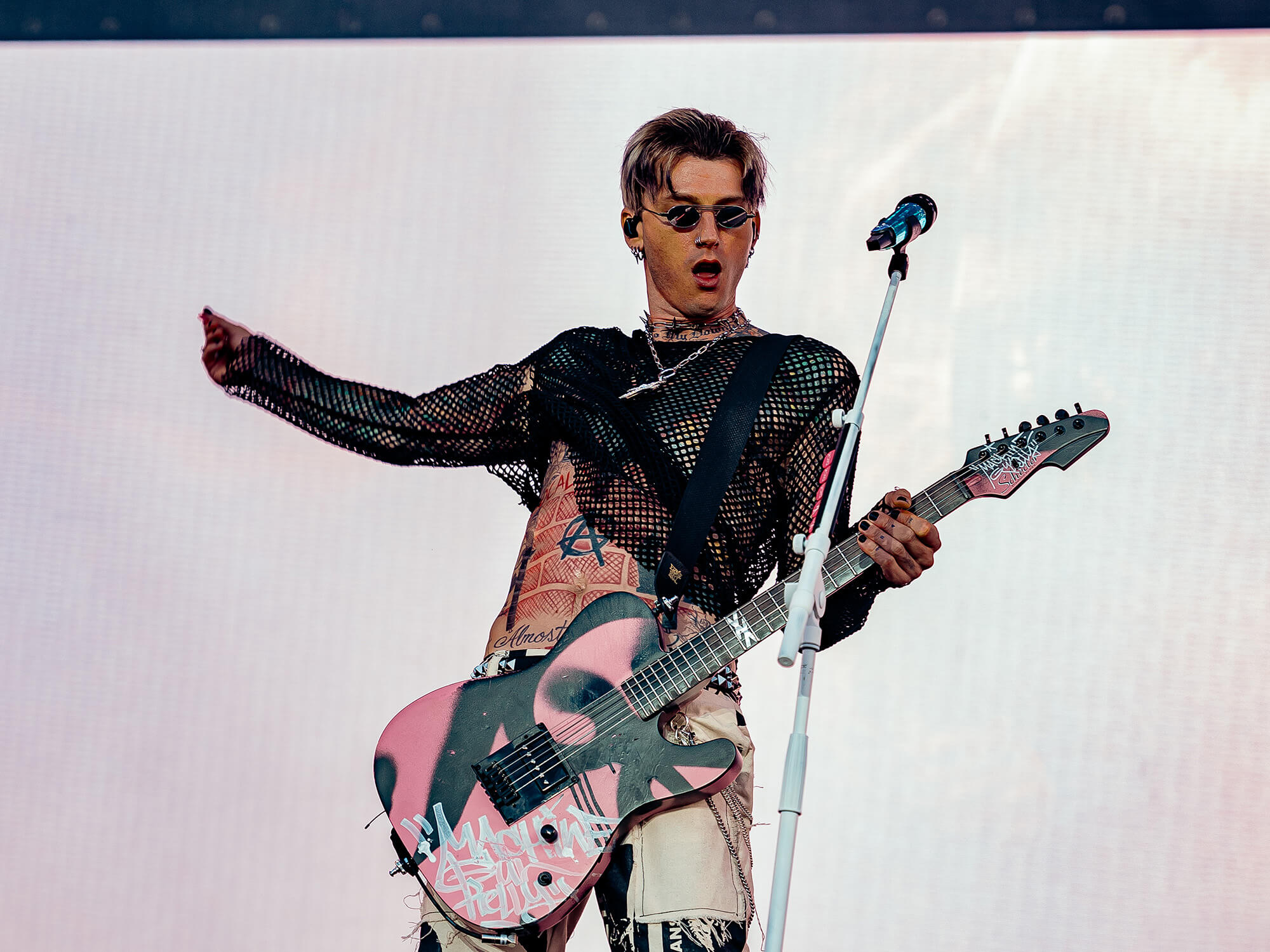 Machine Gun Kelly on stage playing his guitar. It is pink with black spray paint decorating the body. He has one arm out to the side after strumming the instrument.