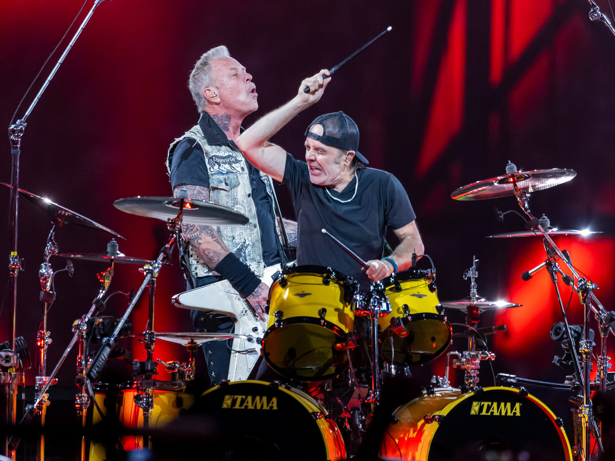James Hetfield and drummer Lars Ulrich on stage. James is playing his white Flying V and Lars has one hand mid-air ready to hit a drum.