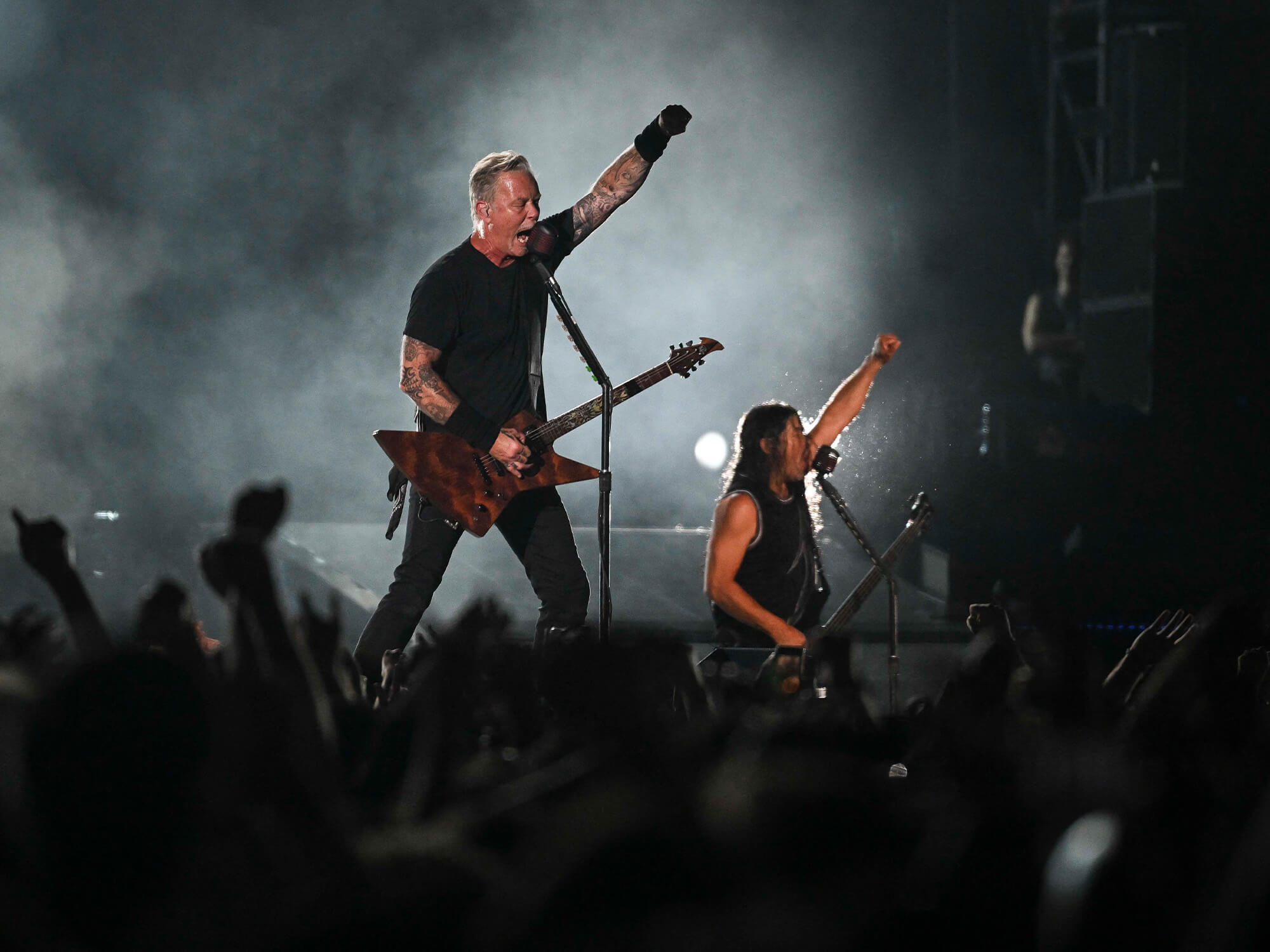 James Hetfield and Robert Trujillo on stage. Both hold on hand to the air and a dark, shadowy crowd is seen below them.