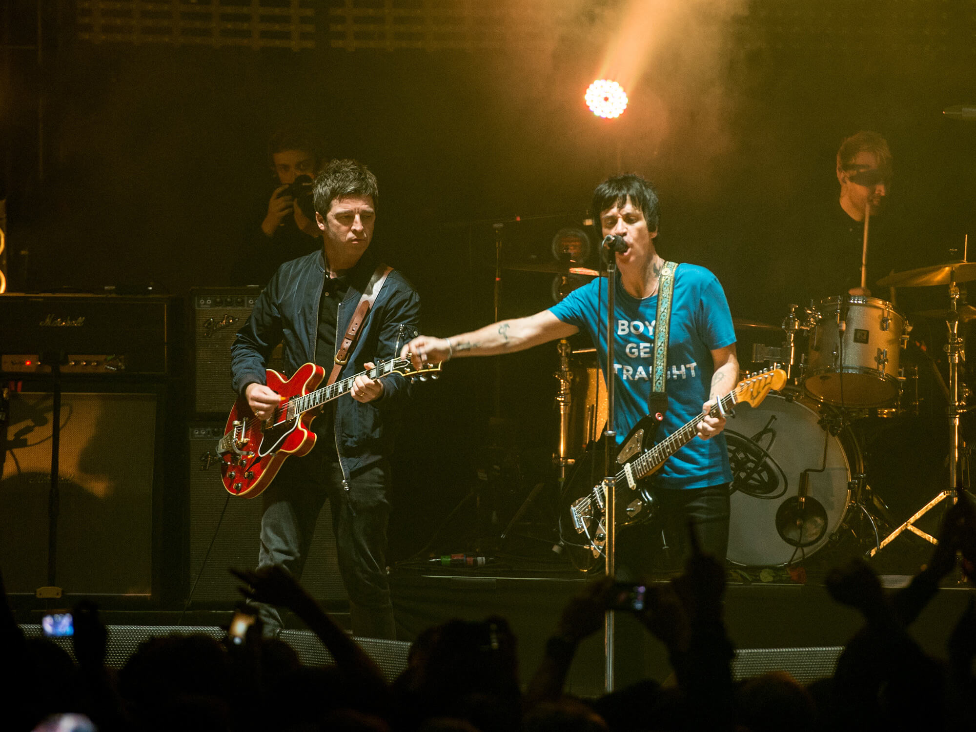 Noel Gallagher and Johnny Marr on stage together in 2014, both with guitars in hand. Marr is singing into a mic and is pointing outwards with his right arm.