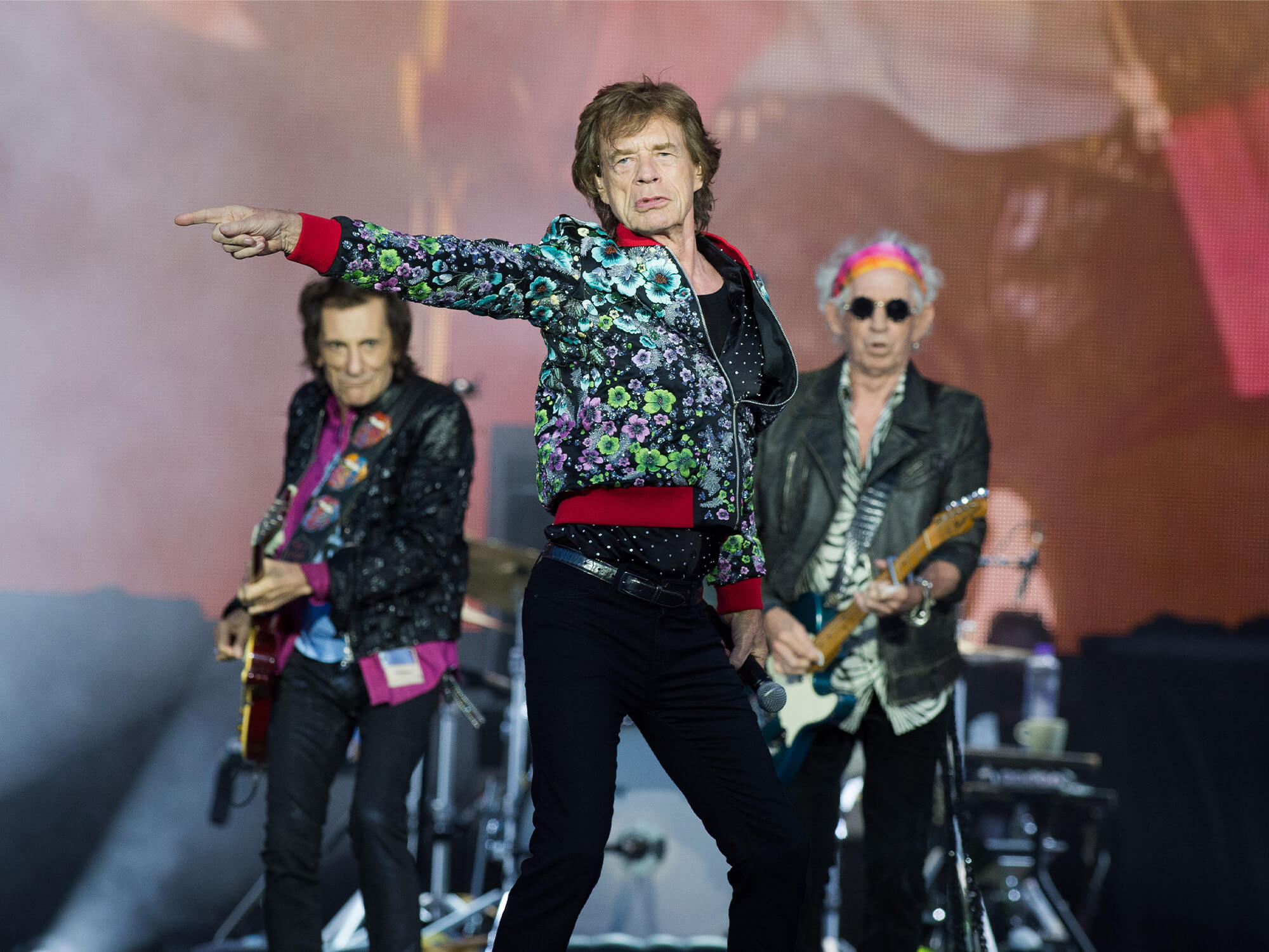 Rolling Stones on stage. Mick Jagger is standing in the middle and pointing to his right. Kieth Richards and Ronnie Wood stand behind him.