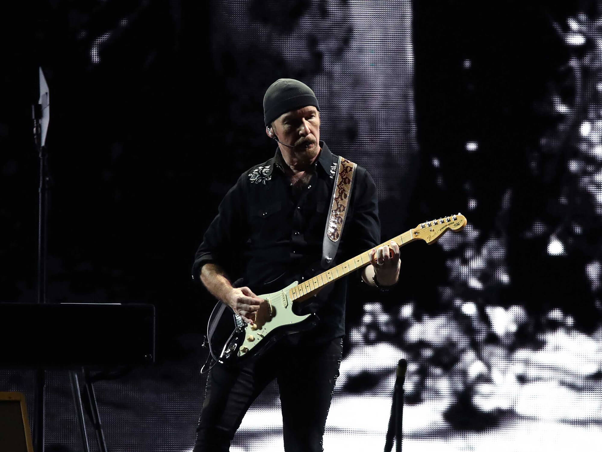 The Edge playing a Stratocaster on stage. There is a small amount of lighting shining on him and the background is dark.