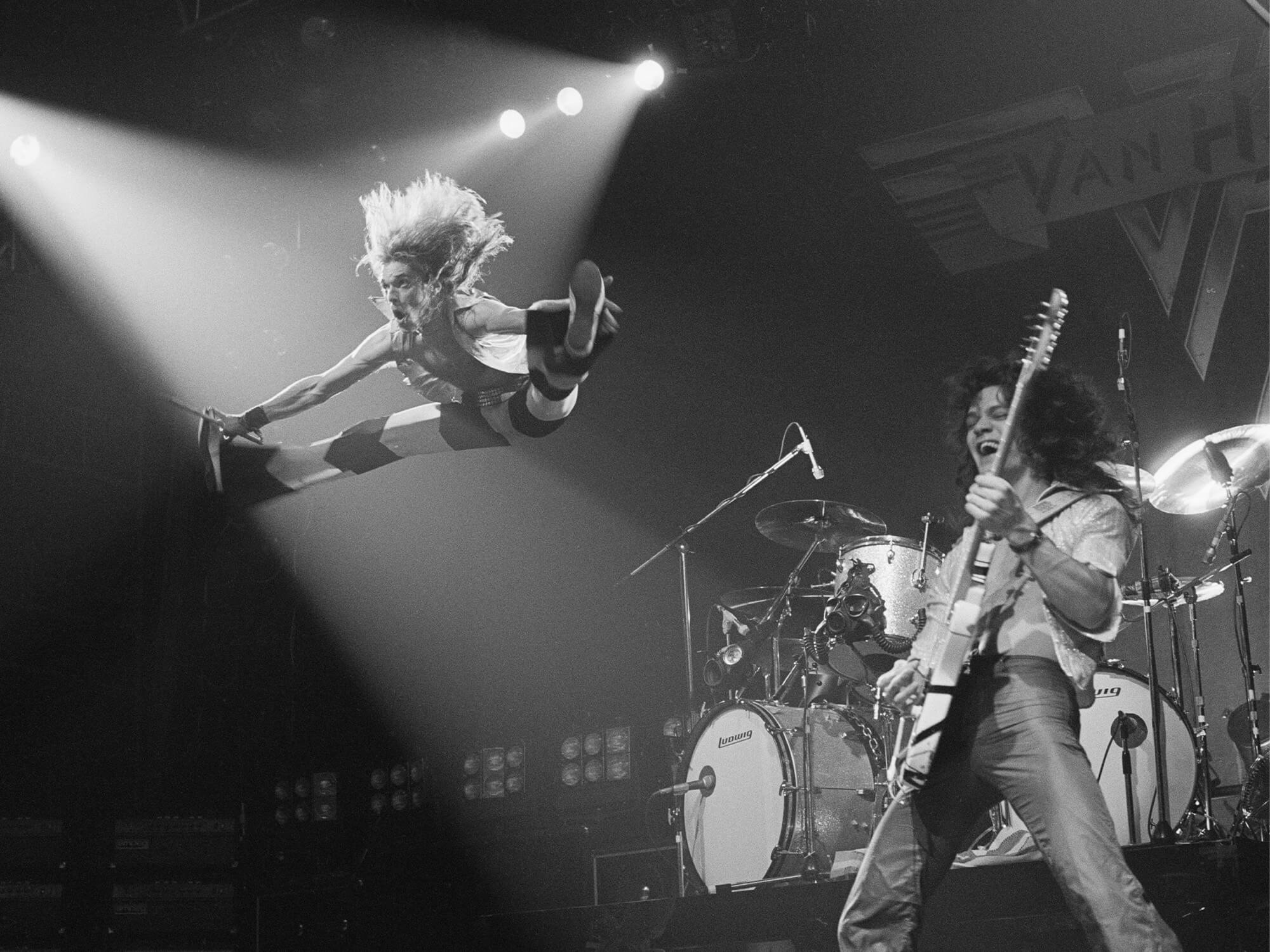 Black and white photo of Van Halen on stage in 1978. David lee Roth is jumping high in the air in the splits, whilst Eddie Van Halen is playing guitar and smiling.
