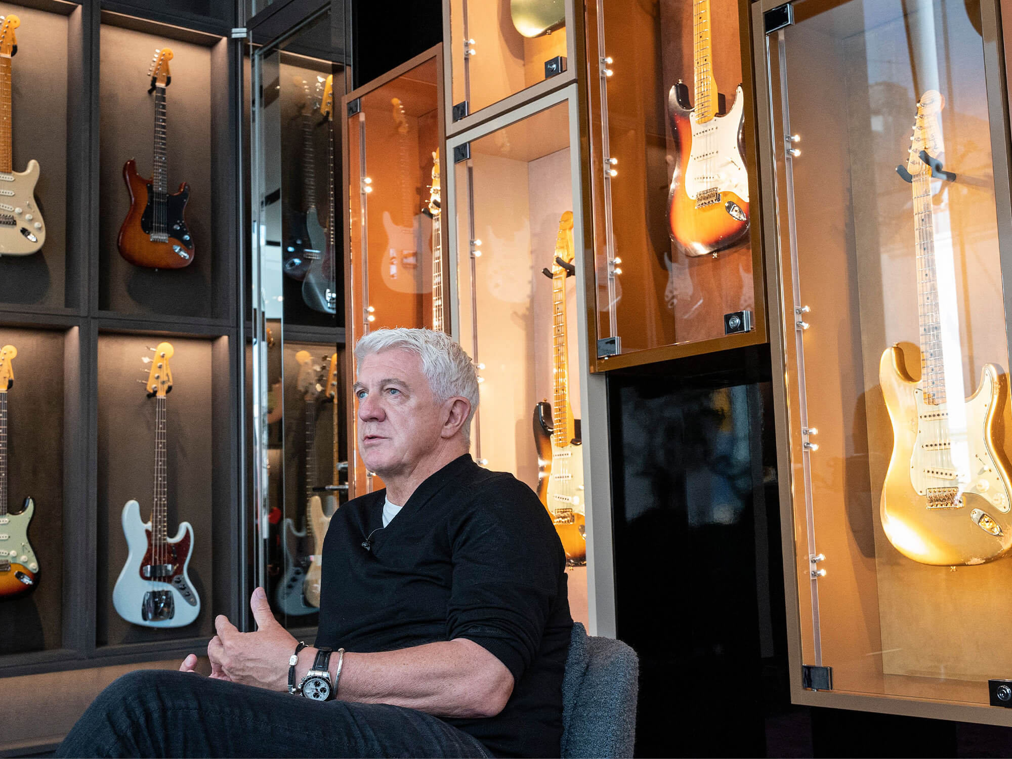Andy Mooney sitting in a chair in front of glass cases filled with Fender guitars. He is engaged in conversation with somebody not in the shot. He has white hair and wears a dark coloured top.