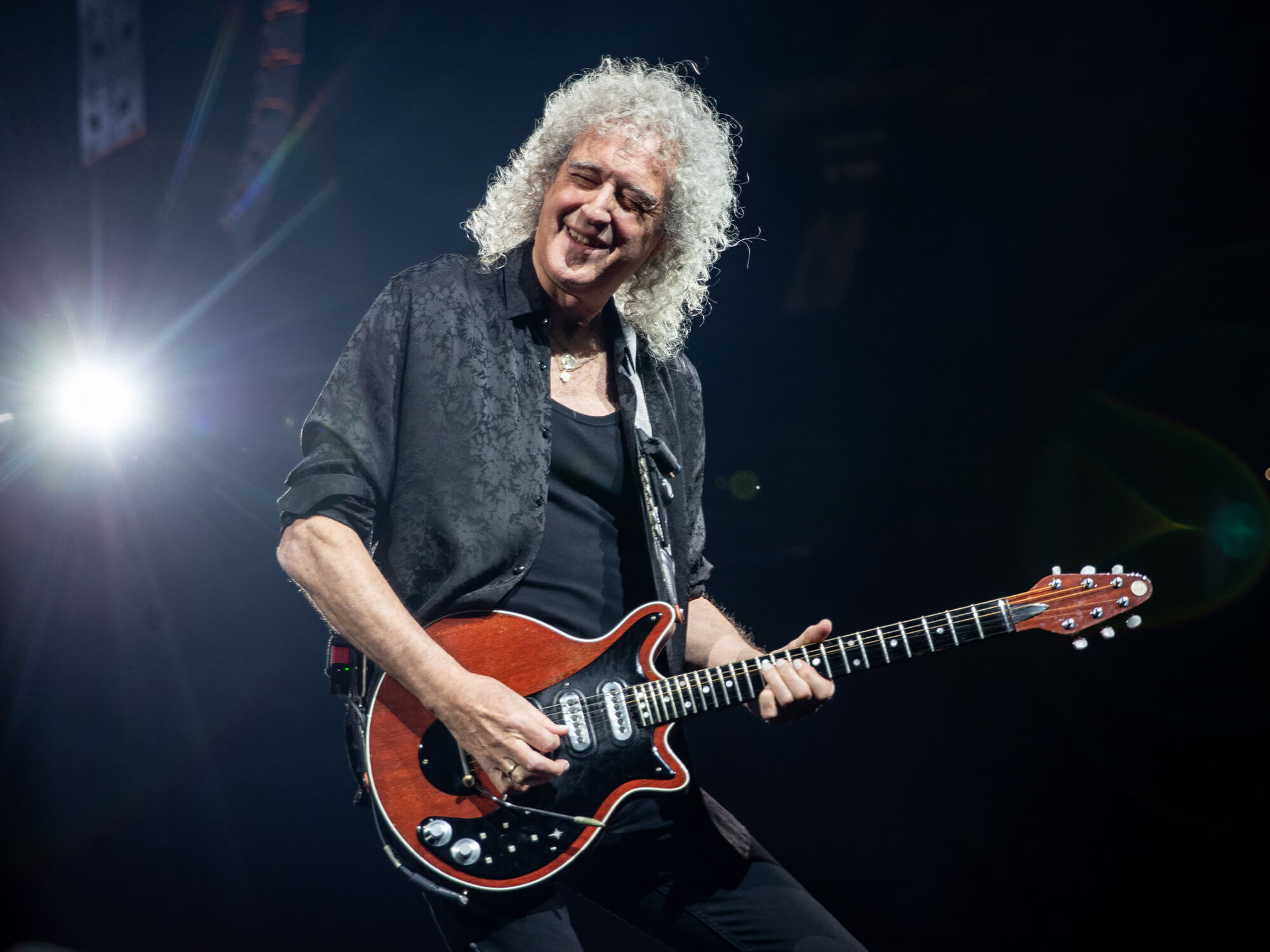 Brian May on stage. He is playing his Red Special guitar and is smiling. He has long curly grey hair and is dressed in black.