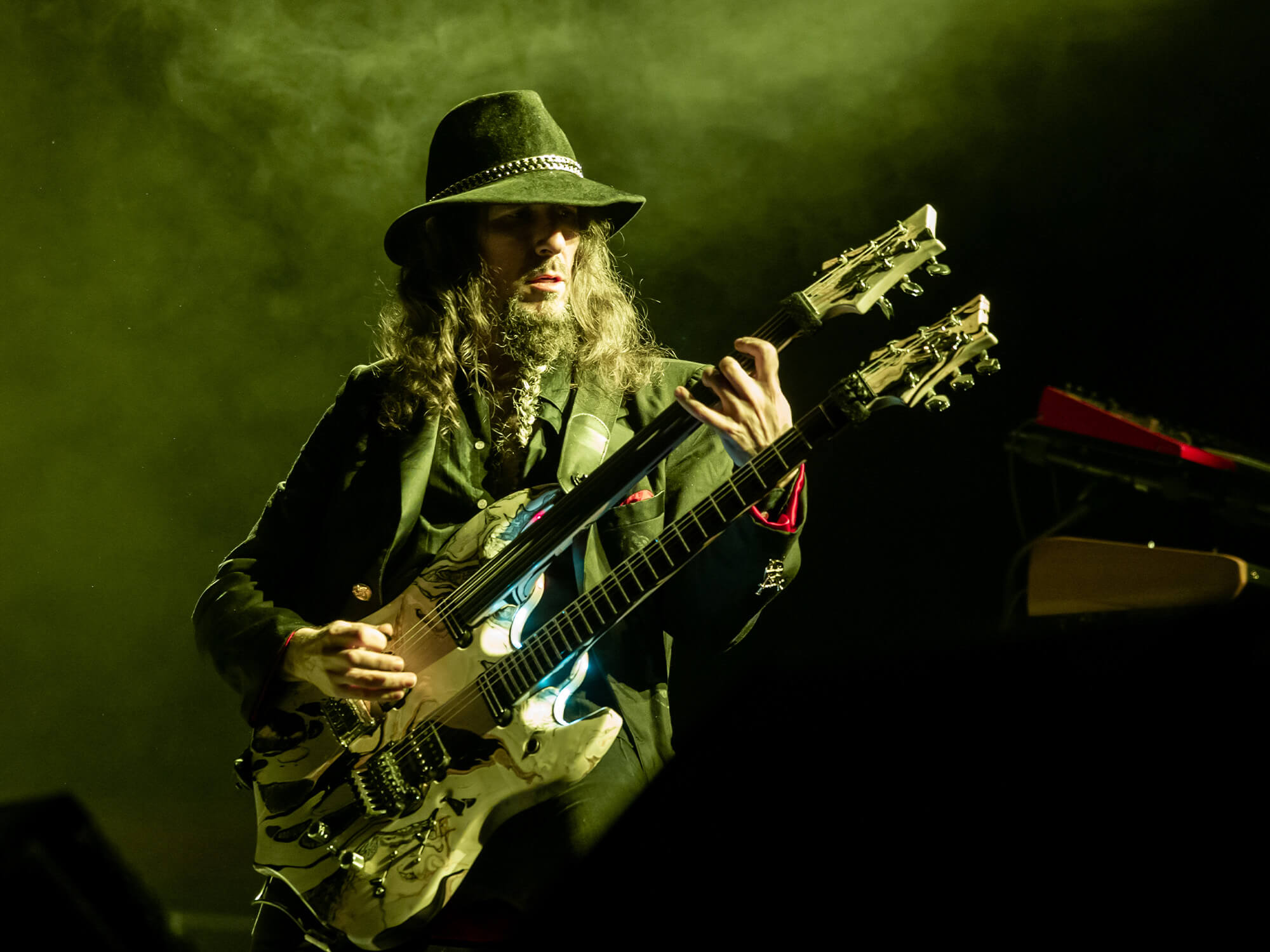 Bumblefoot playing a double neck guitar. He has long hair and is wearing a brown hat. He stands under green lighting.