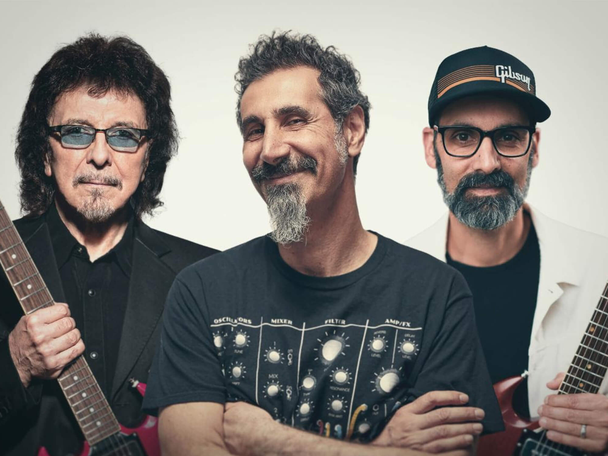 Tony Iommi, Serj Tankian, and Cesar Gueikian. Serj is smiling with his arms folded, while Cesar and Tony are holding their guitars behind him.