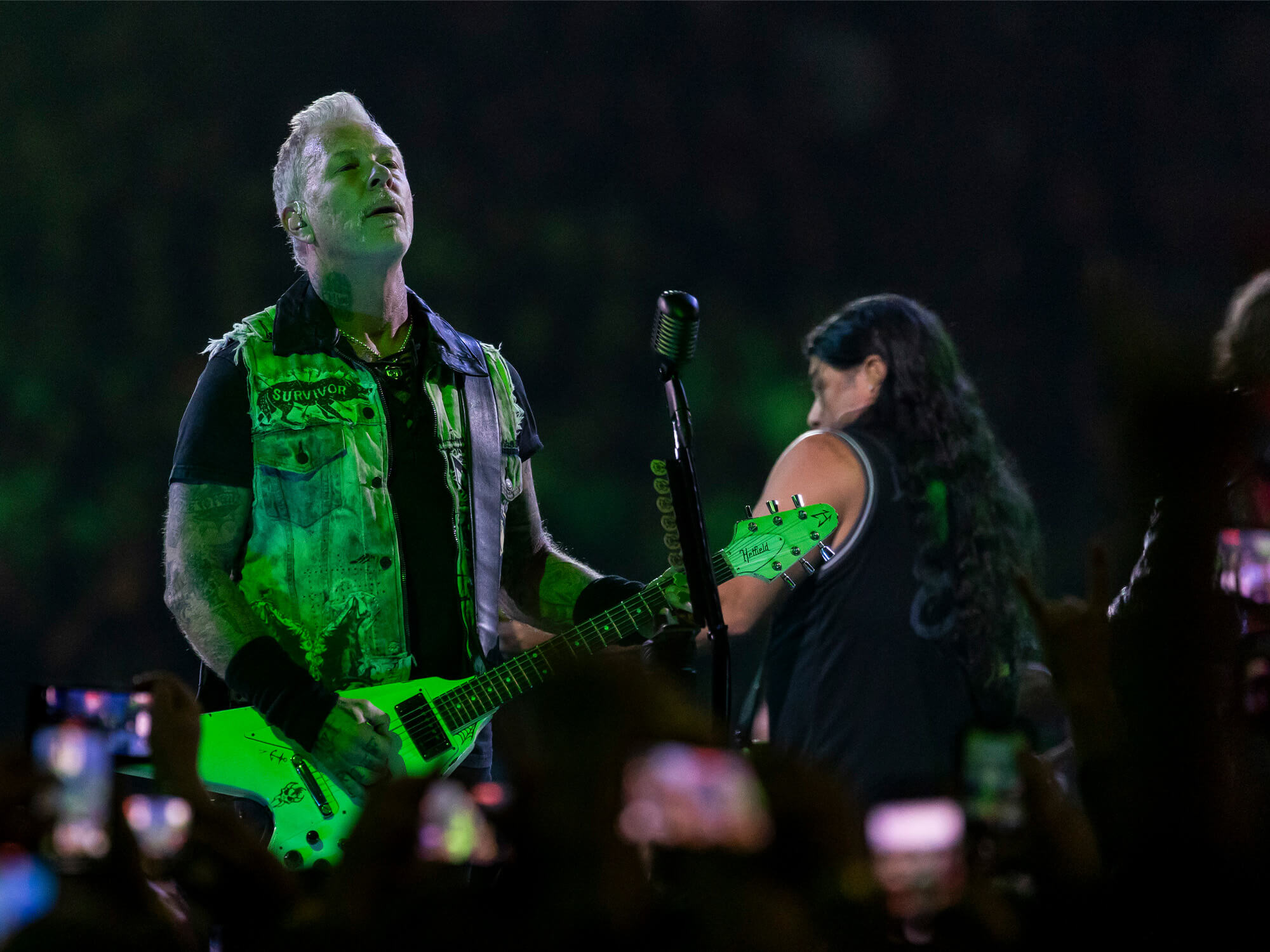 James Hetfield on stage playing guitar. He is standing by bassist Robert and is looking up above him. His face and body is illuminated by green lights.