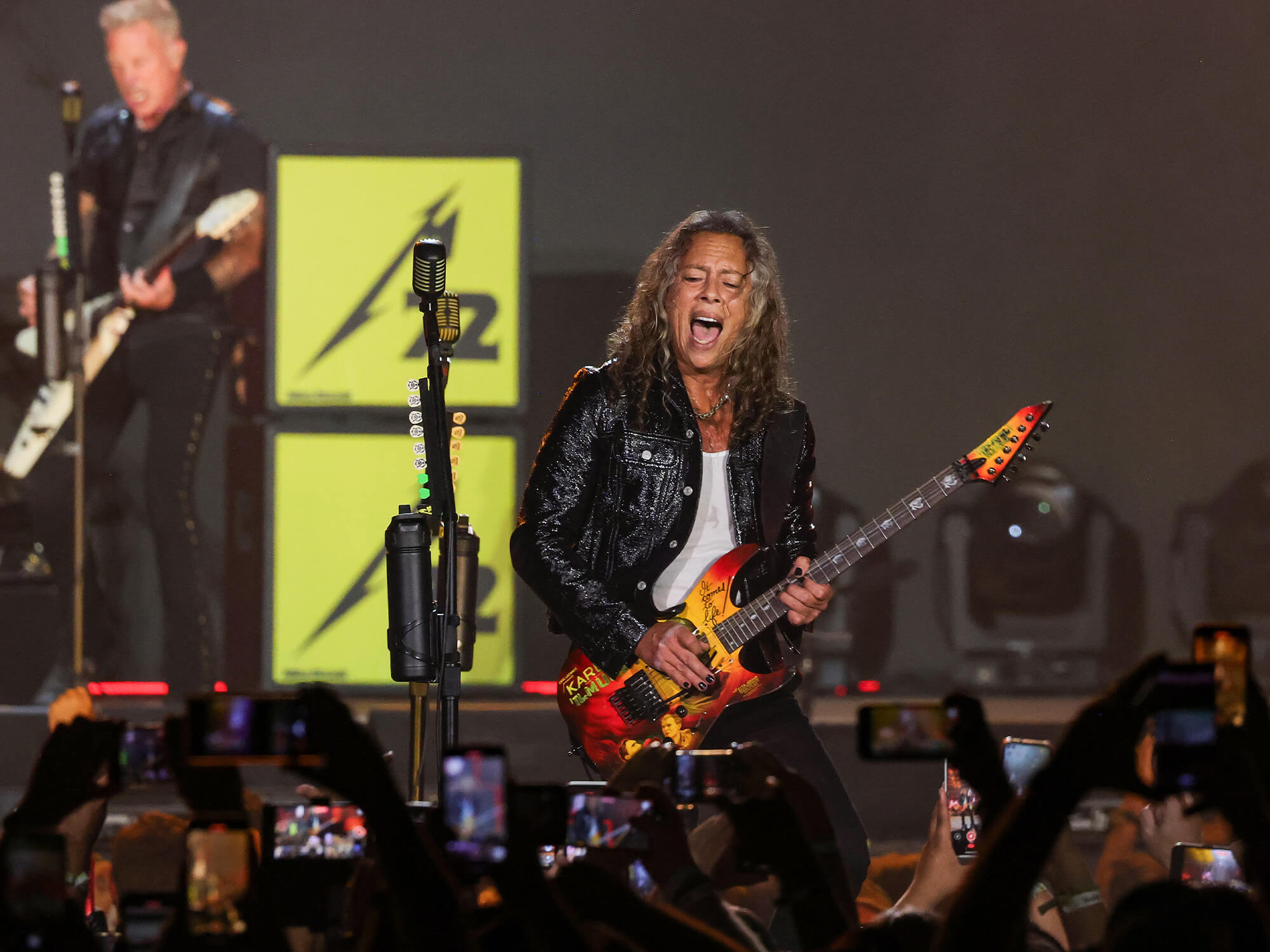 Kirk Hammett playing guitar. He has his mouth open as if shouting or singing, and has long hair and dark clothes on.