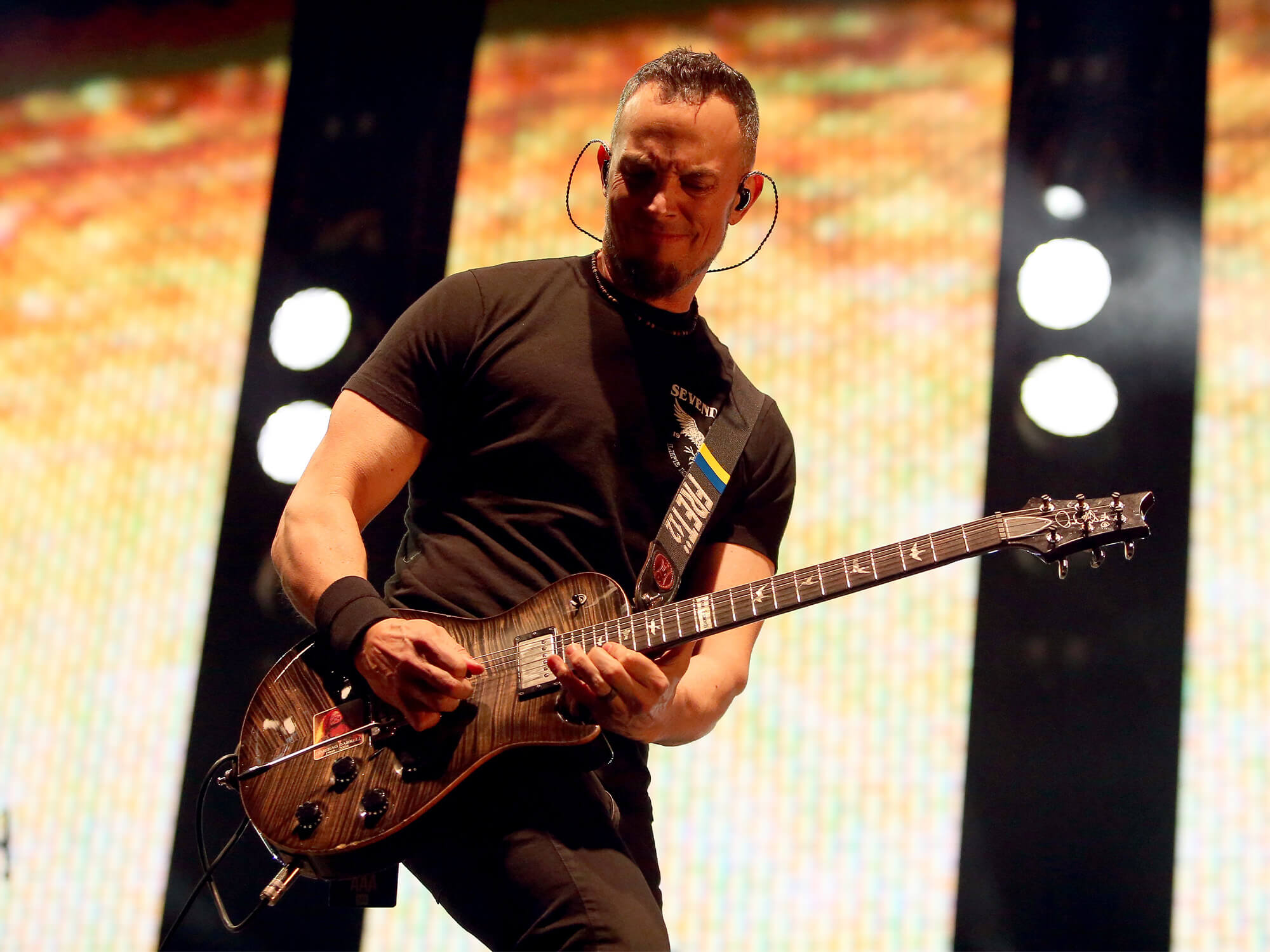 Mark Tremonti on stage playing guitar. He has a grey-coloured PRS model and is wearing all black. He is playing high up on the fretboard close to the body.