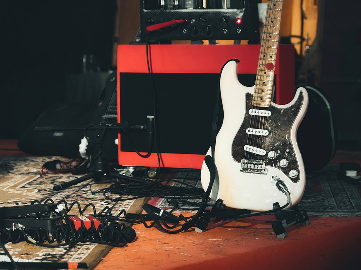 A white electric guitar and a red amplifier onstage surrounded by cables and effects pedals, photo by Tennessee Witney/Getty Images