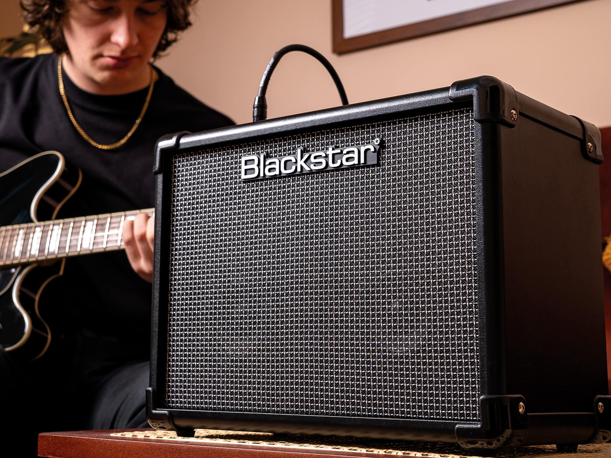 Blackstar ID:CORE V4 amplifier in use. A person is sitting behind the amp and is playing guitar. The amplifier is black all over, small in size, and features the Blackstar logo on the grille.
