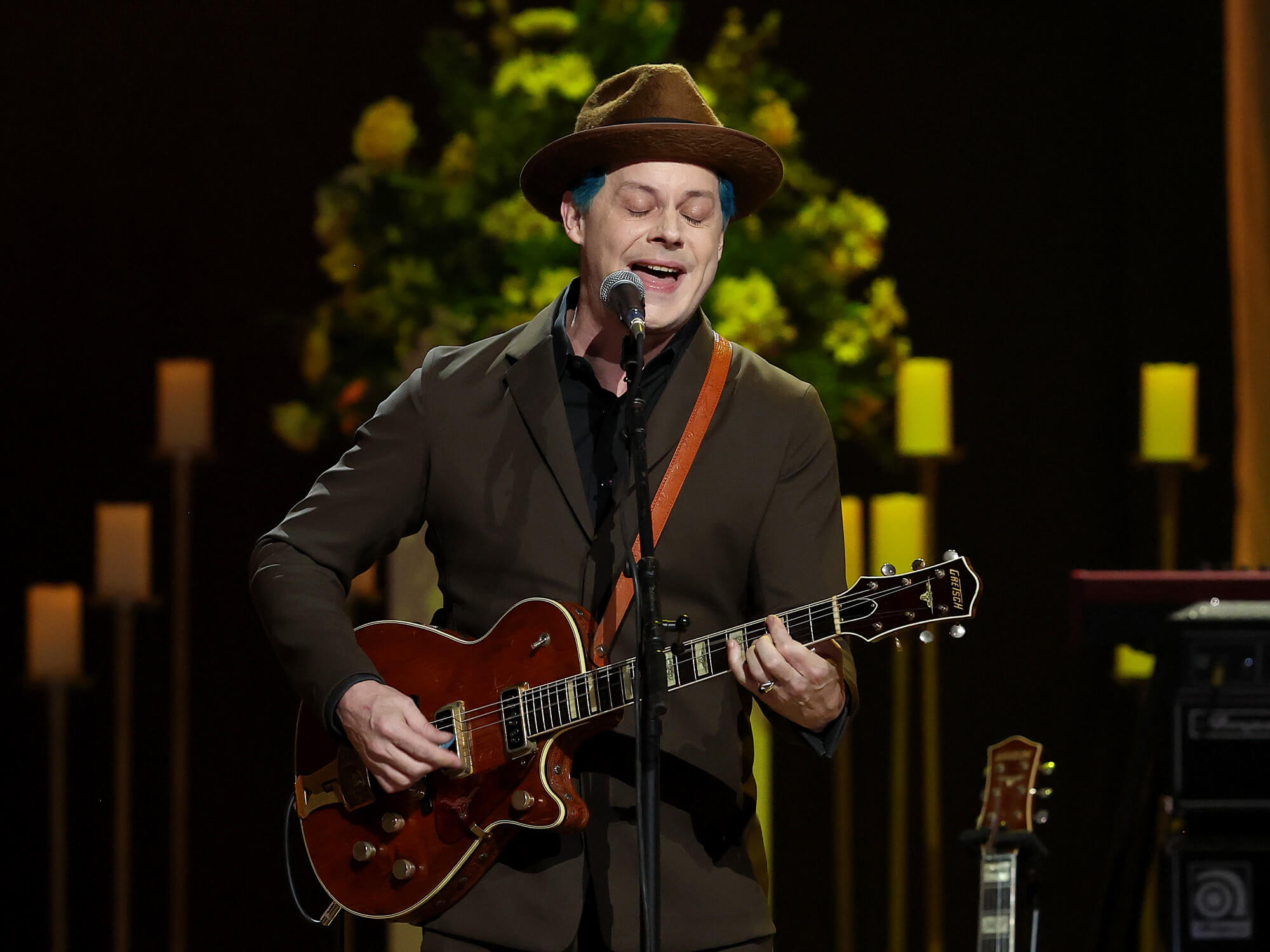 Jack White on stage. He is singing into a mic on a stand and is playing a Gretsch electric guitar. He wears a brown hat and suit jacket. His bright blue hair can be seen just peeping from underneath the hat.