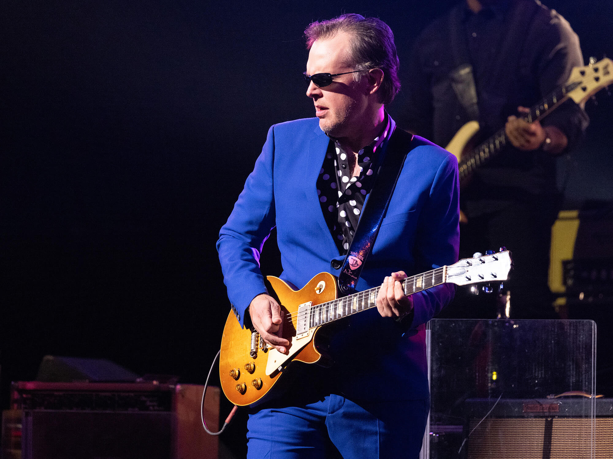 Joe Bonamassa on stage. He is wearing a bright blue suit and sunglasses, and is playing a Les Paul guitar.