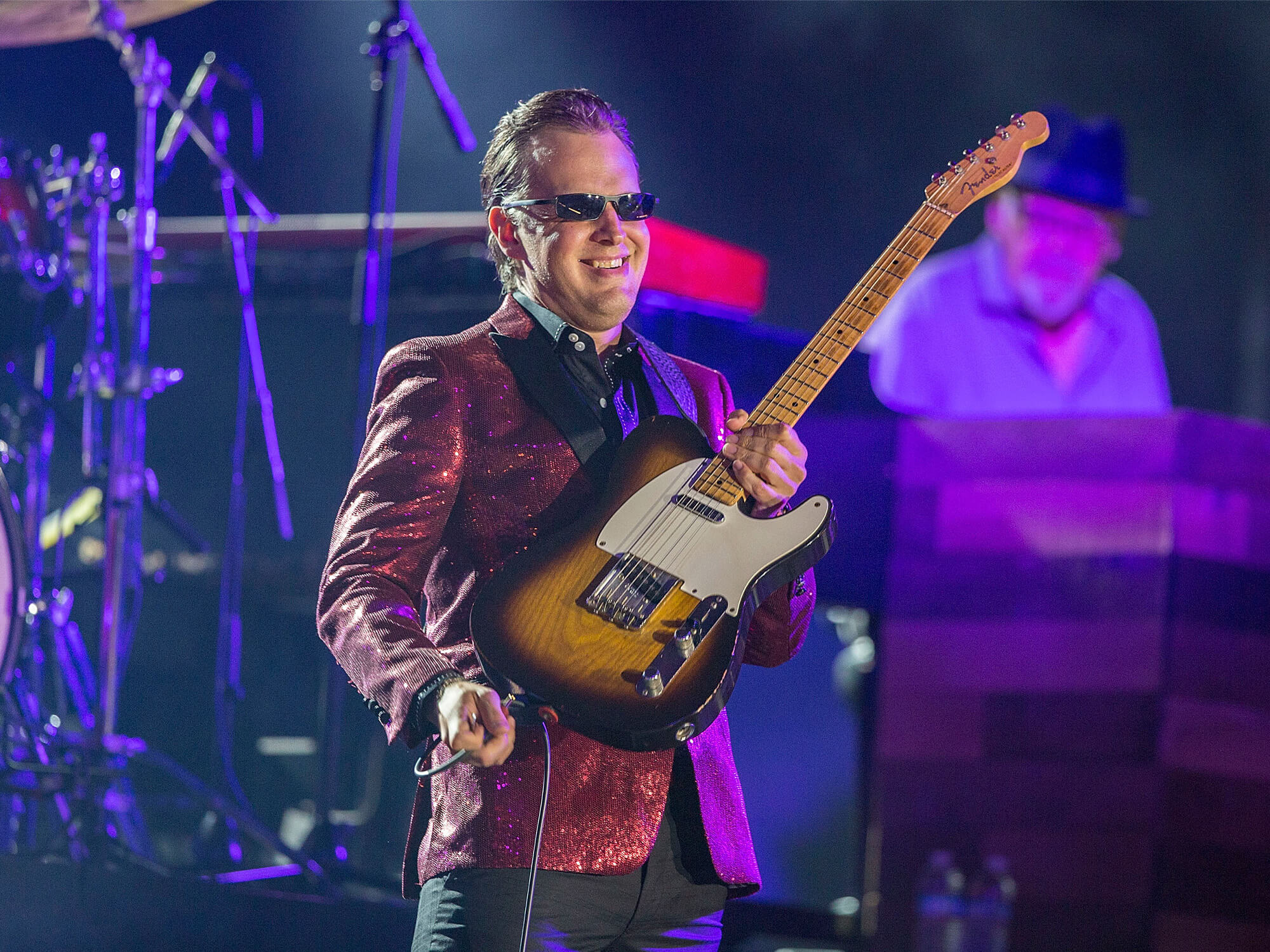 Joe Bonamassa on stage. He is holding up a Telecaster guitar and smiling. He is wearing a red suit jacket and blacked-out sunglasses.